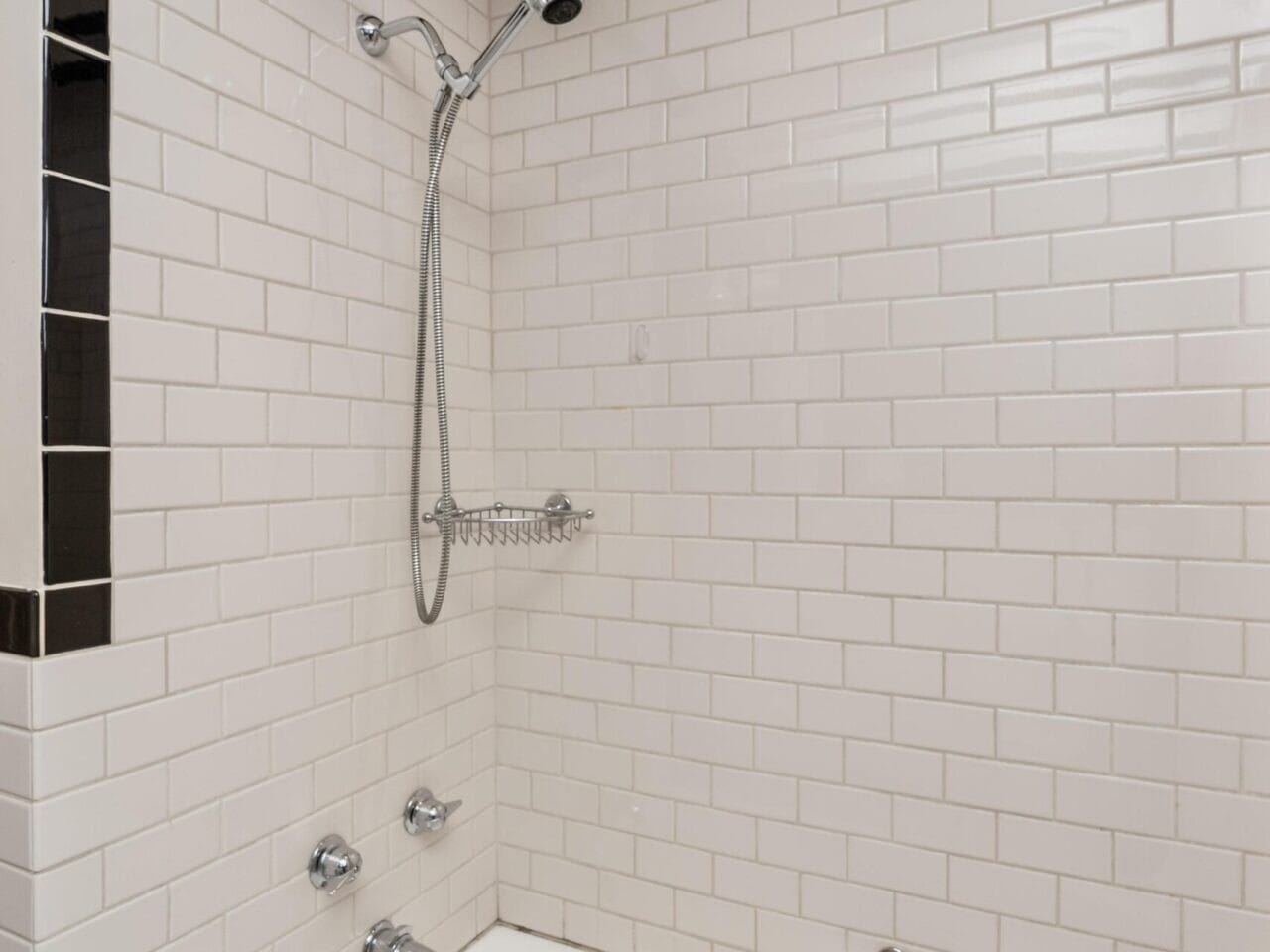 A bathroom in a Portland, Oregon real estate gem features a white tile shower with black trim and a wall-mounted showerhead. It includes a built-in soap holder and bathtub below. Soft lighting enhances the space, complete with a small wall-mounted shelf for toiletries, curated by your top Portland realtor.