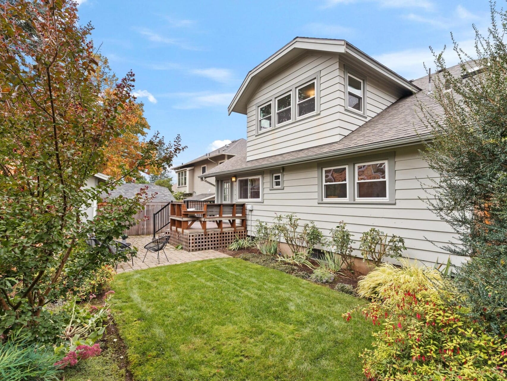 A two-story house with light siding and a small patio set in a fenced yard. The garden features green grass, shrubs, and a tree with autumn leaves. The sky is blue with scattered clouds.