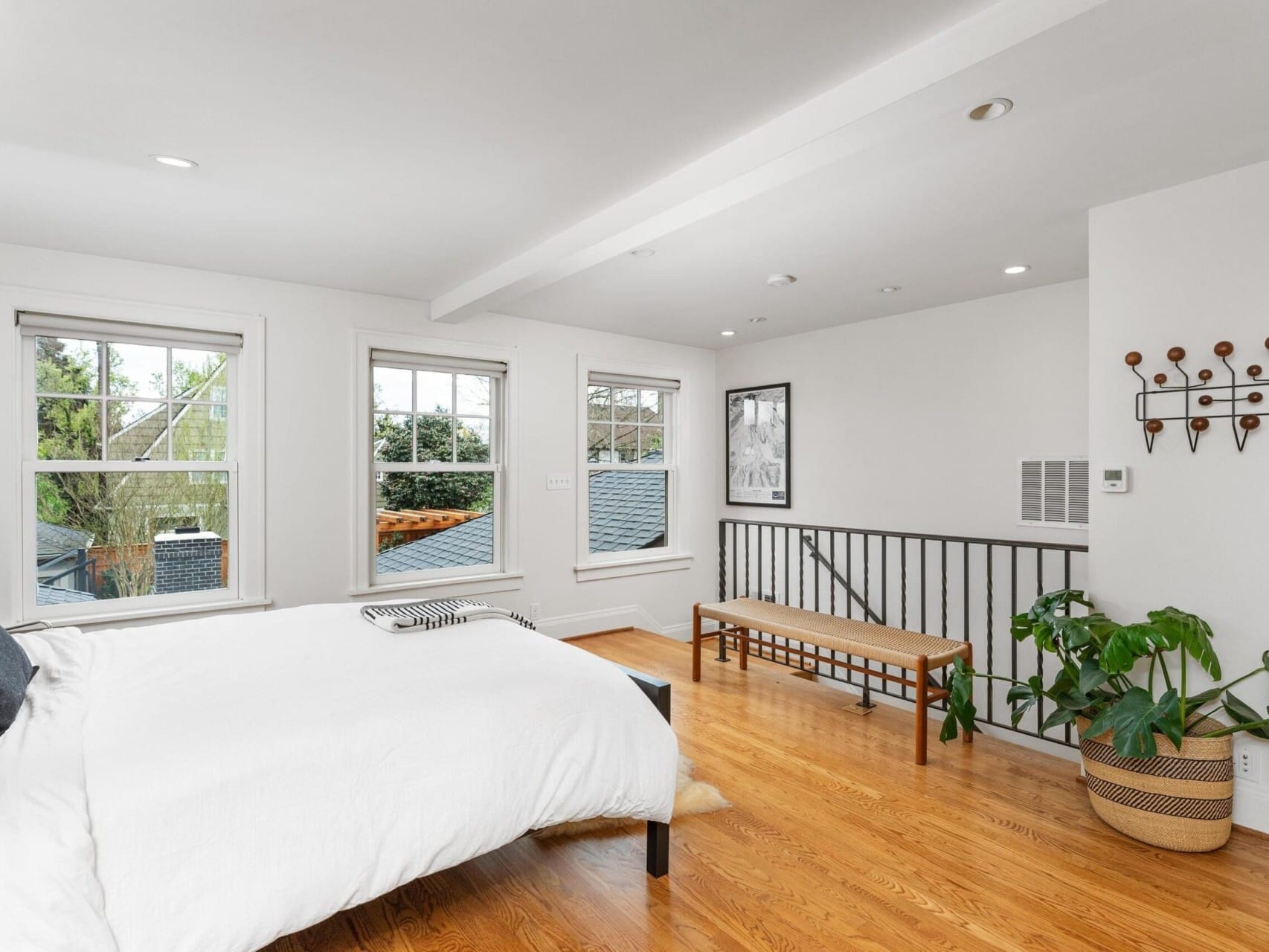 A bright bedroom with a large bed, white linens, and wood flooring awaits in this charming Portland Oregon real estate find. Three windows allow natural light, while a potted plant and wooden bench add warmth. Perfectly presented by your top real estate agent in town, it’s a cozy haven you'll love.