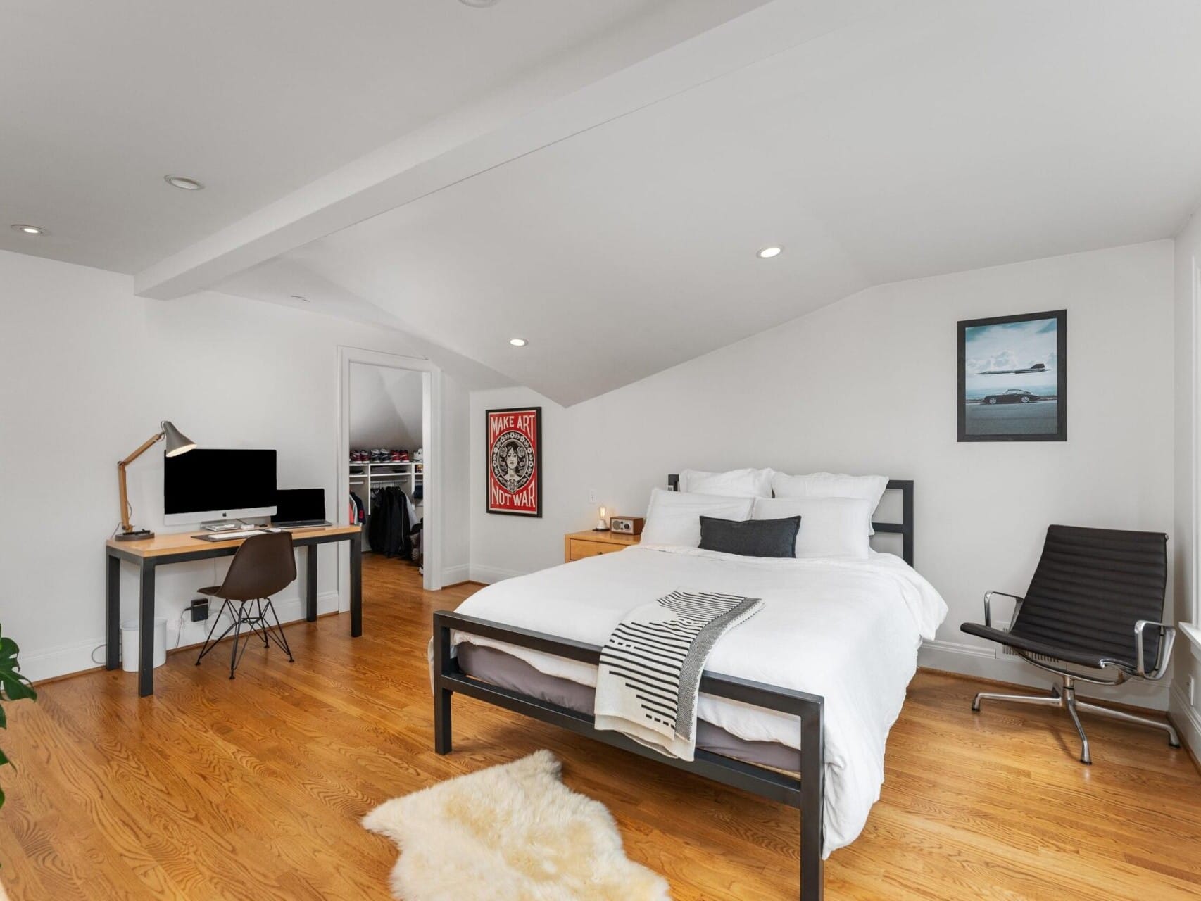 A modern bedroom in Portland Oregon real estate features a sloped ceiling, a neatly made bed, a black office chair, and a wooden desk with a computer. Natural light floods in through large windows, highlighting hardwood floors and minimalistic decor, including a framed poster.