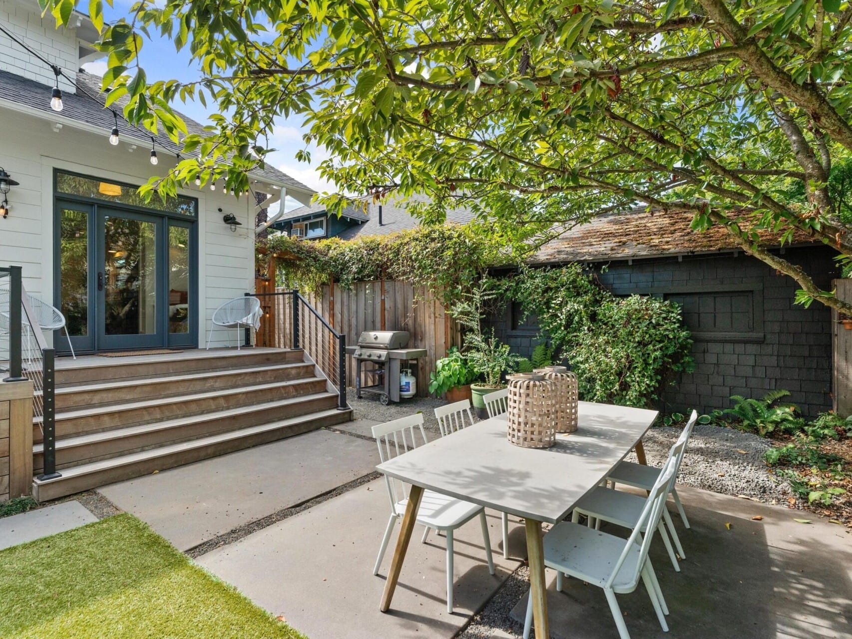 A cozy Portland, Oregon backyard with a modern patio. Features a white house with steps leading to a glass door, a wooden dining table with white chairs, a barbecue grill, string lights above, and lush greenery surrounding the area.