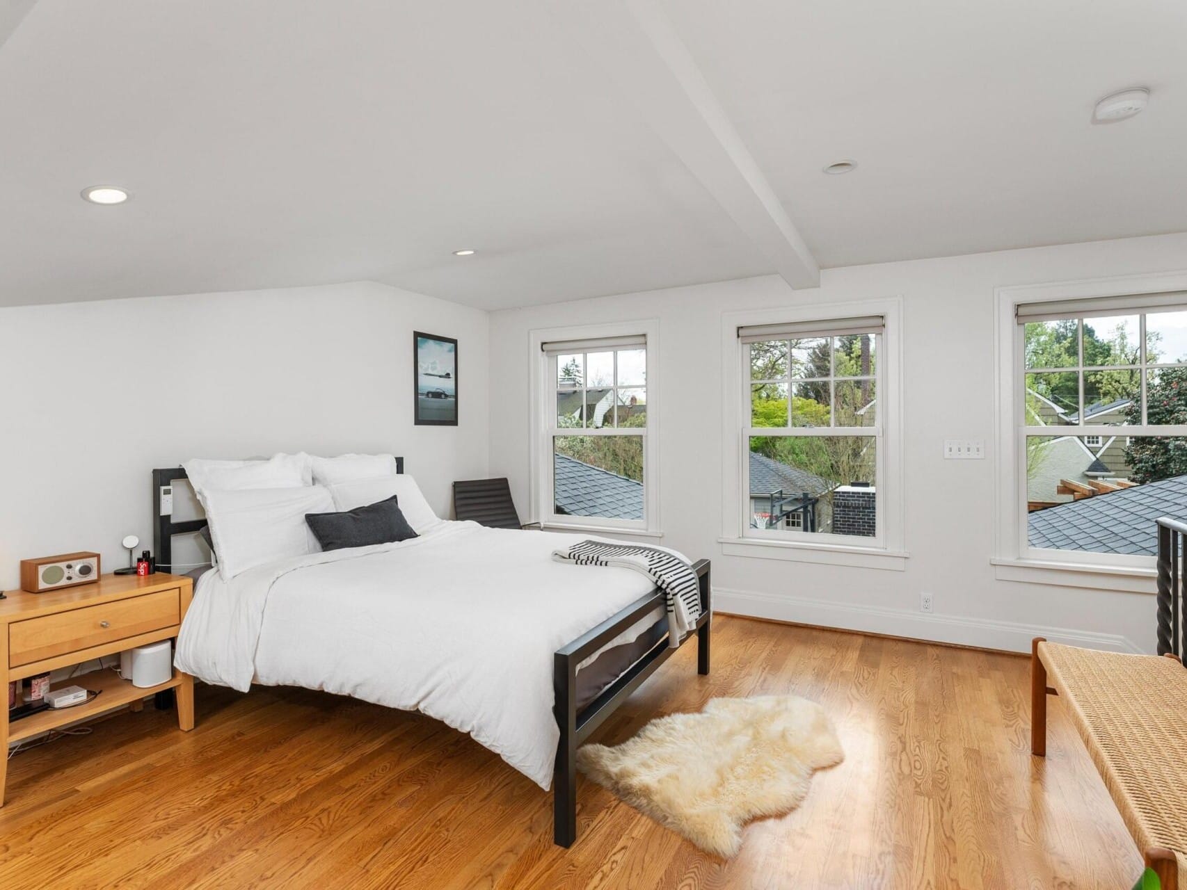 This Portland Oregon real estate gem features a bright bedroom with a large bed, white bedding, and two pillows. A wooden bedside table with a lamp and clock sits to the left. Three windows overlook greenery, complementing the hardwood floors and small rug. A bench is near the railing on the right.