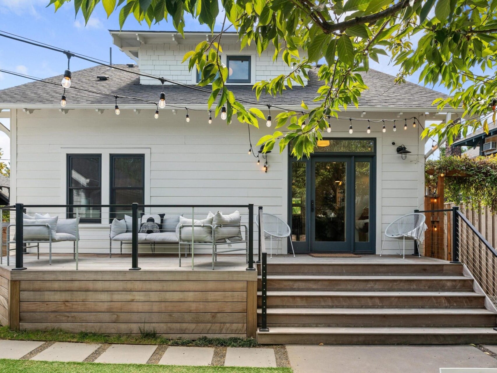 A charming white house in Portland, Oregon, boasts a patio with outdoor seating and string lights. The wood-framed glass doors welcome you inside, while a wooden staircase ascends from a stone path surrounded by lush greenery and a tree in the foreground.