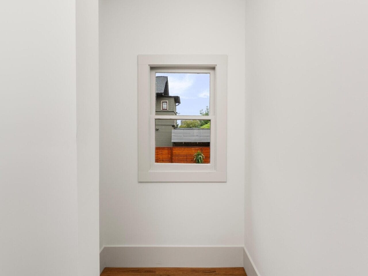 A small, narrow hallway with white walls and a wooden floor leads to a window overlooking a neighboring Portland, Oregon house and a patch of blue sky. A circular ceiling light gently illuminates the space.