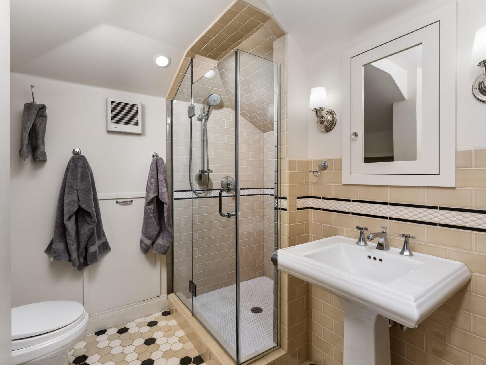 A compact bathroom in a Portland Oregon real estate listing boasts a glass-enclosed shower with beige tiles, a white pedestal sink, and a wall-mounted mirror. Hexagonal floor tiles are black, white, and beige, while gray towels hang nearby. Small wall lamps accentuate the space's elegant charm.