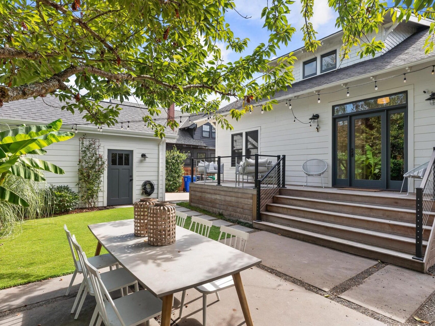 A sunny backyard in Portland, Oregon features a patio area with a white dining table and chairs. Steps lead to a house adorned with string lights above. A tree provides shade amid the lush greenery of plants and bushes. A separate garage is also visible.