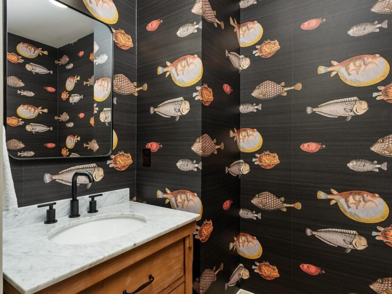 A small bathroom in Portland, Oregon, features fish-patterned wallpaper. It includes a wooden vanity with a marble top, a black faucet, a rectangular mirror, and a wall-mounted light with a white shade. The floor is light wood, adding to its cozy charm.