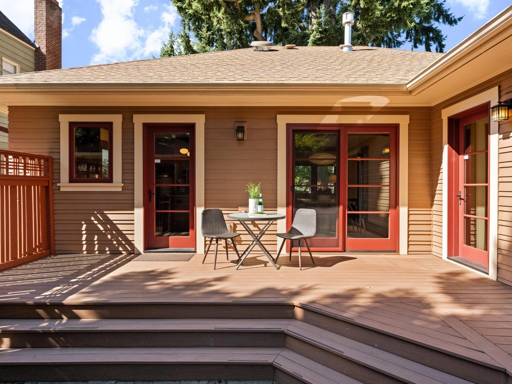 A wooden deck in Portland, Oregon features a small round table and two chairs. The deck connects to a house with charming red-framed doors and windows. A potted plant adorns the table, while sunlight dances and trees stand tall in the background.