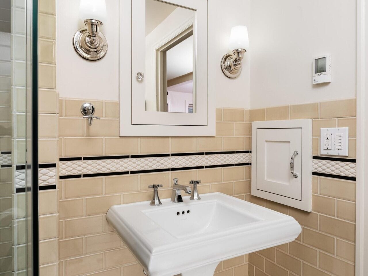 A small bathroom corner in a Portland Oregon real estate gem features a white pedestal sink below a mirrored cabinet. The tan tiled walls have a black and white border, and two wall-mounted lights are on either side of the mirror. Hexagonal floor tiles complete the classic look.