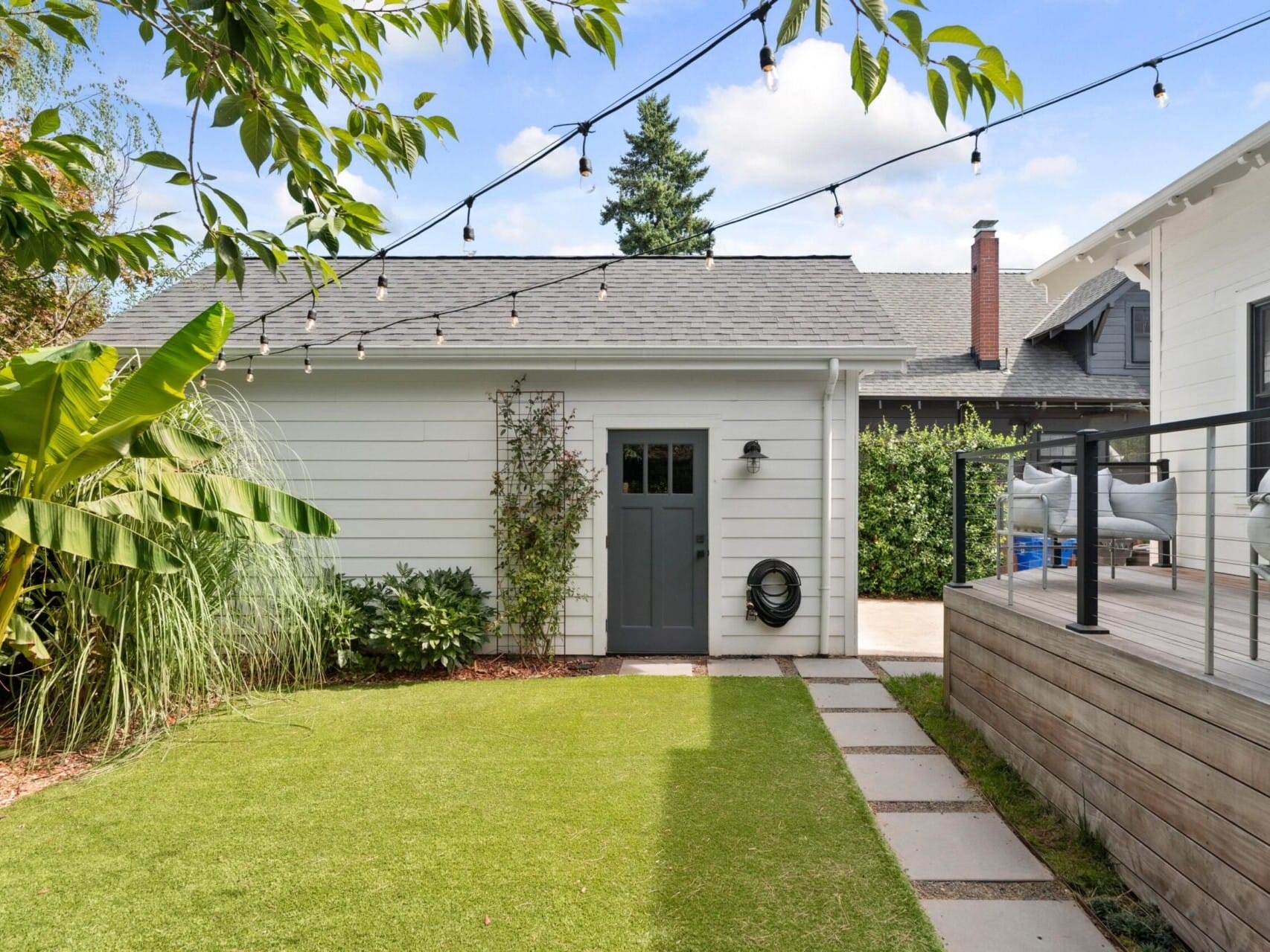 A Portland, Oregon backyard featuring a small white shed with a dark door, surrounded by lush greenery. String lights twinkle overhead, illuminating a deck on the right with cozy outdoor seating. The neatly landscaped lawn is adorned with stepping stones leading to the inviting shed.