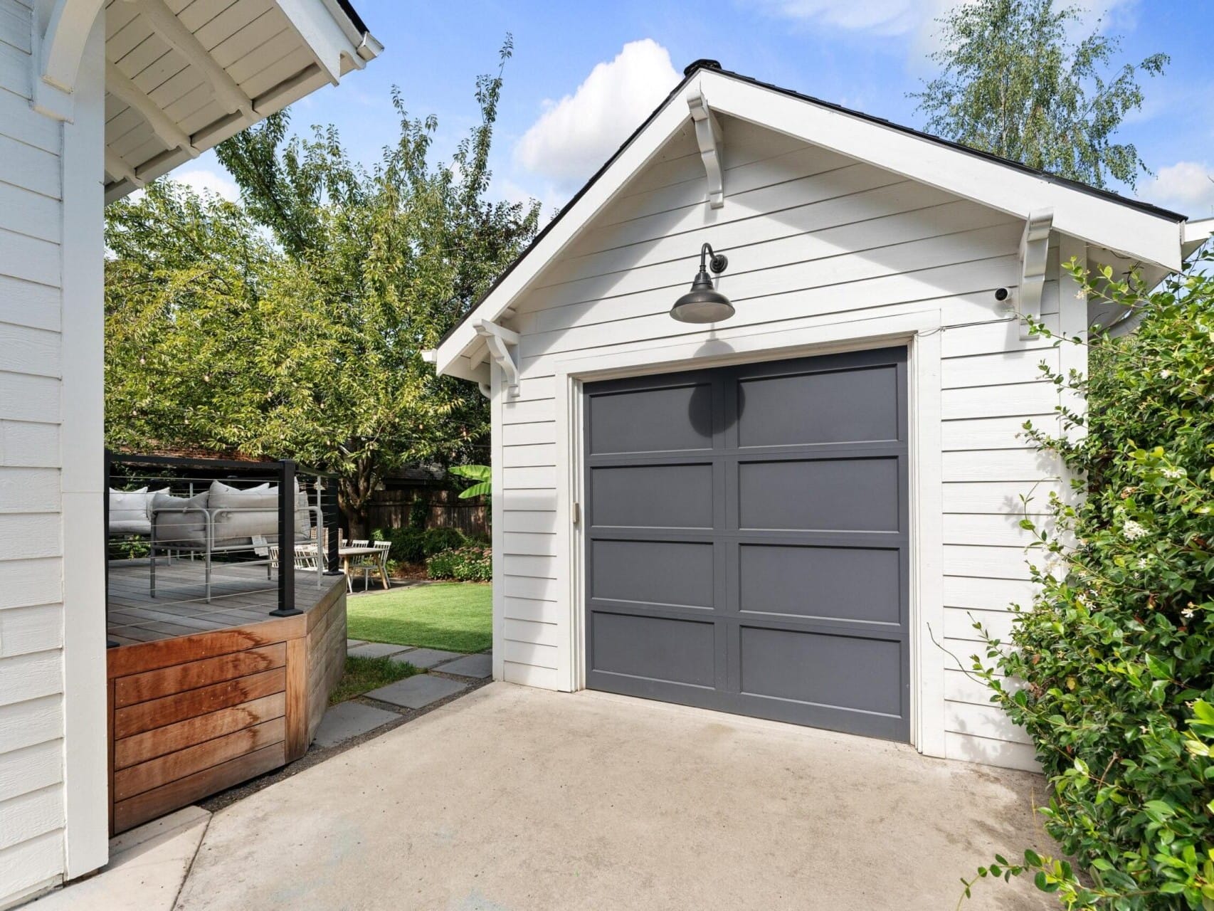 A light gray garage with a dark gray door is surrounded by lush Portland, Oregon greenery. There's a wooden deck to the left with outdoor seating and a table. A tree provides shade, while the sky remains clear with a few clouds drifting lazily.