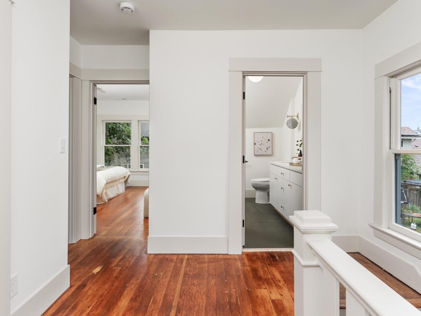 The interior hallway, nestled in a charming Portland, Oregon home, features wooden floors and a white wall. It offers views into a cozy bedroom with large windows and a stylish bathroom with a vanity and toilet. A window on the right frames an inviting outdoor scene.