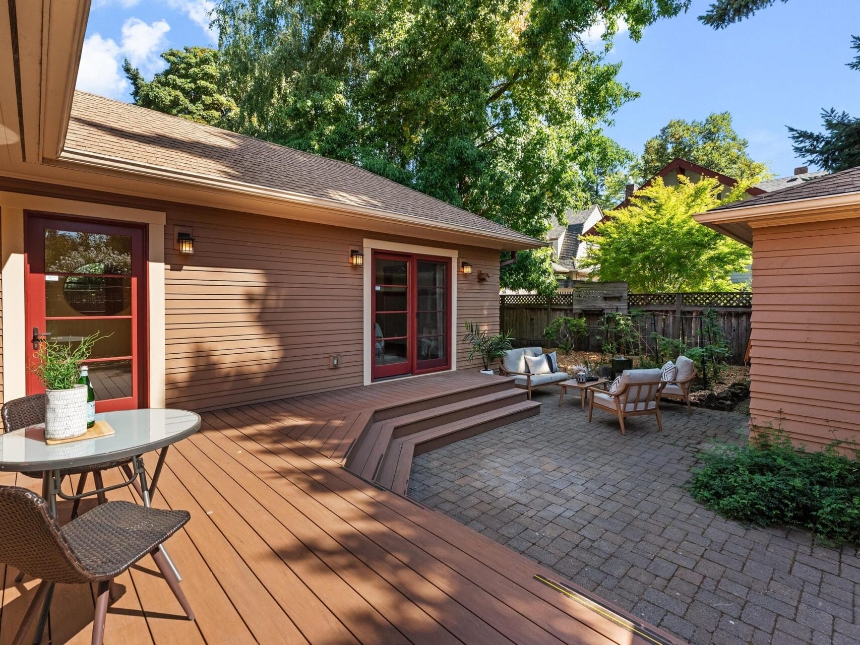 In a serene Portland, Oregon backyard, a wooden deck hosts a small table and chairs. Steps lead to a stone patio with additional seating. Lush greenery and a wooden fence offer privacy, while red doors open to the house and tall trees cast soothing shade.