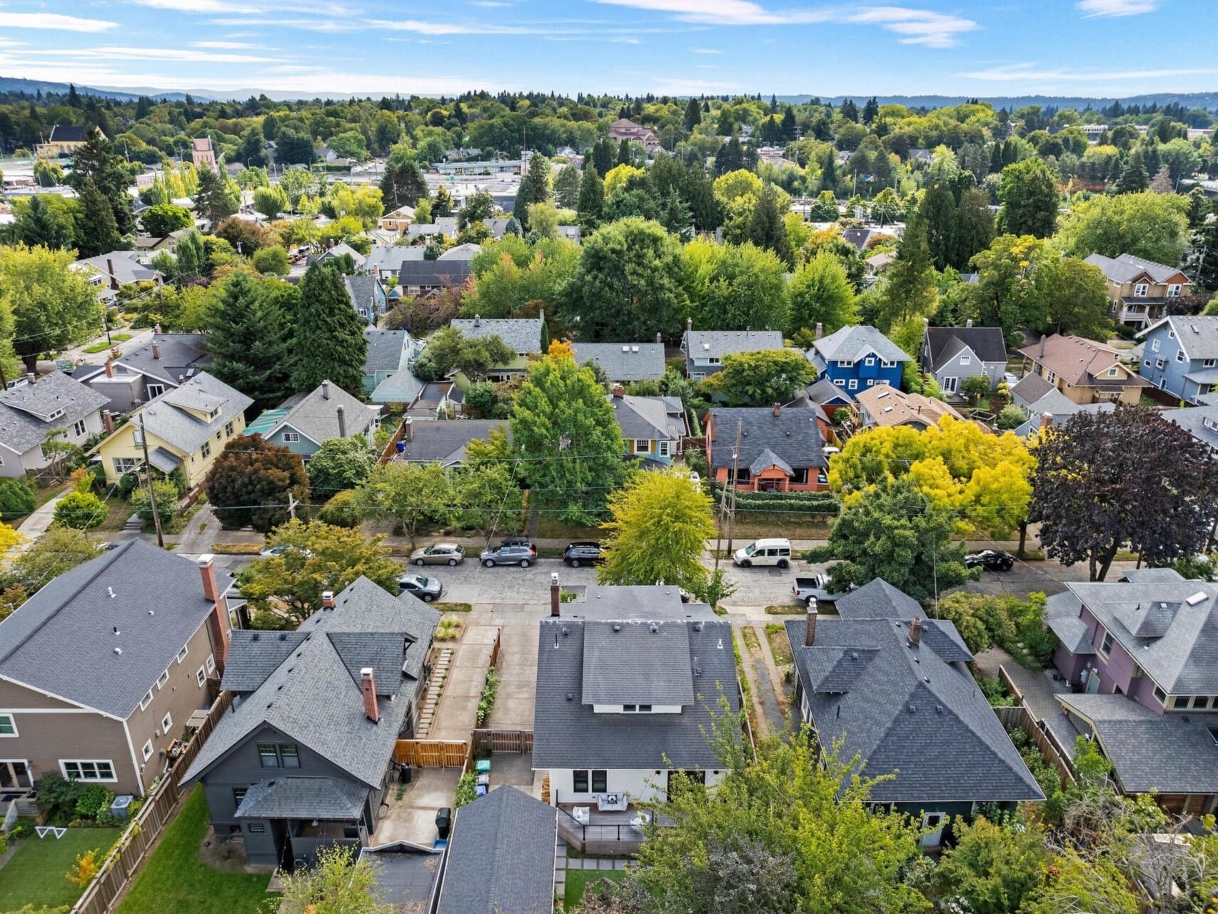 Aerial view of a suburban neighborhood in Portland, Oregon, featuring tree-lined streets and houses with diverse roof colors. The area is lush with greenery, and distant hills can be seen on the horizon under a clear sky.