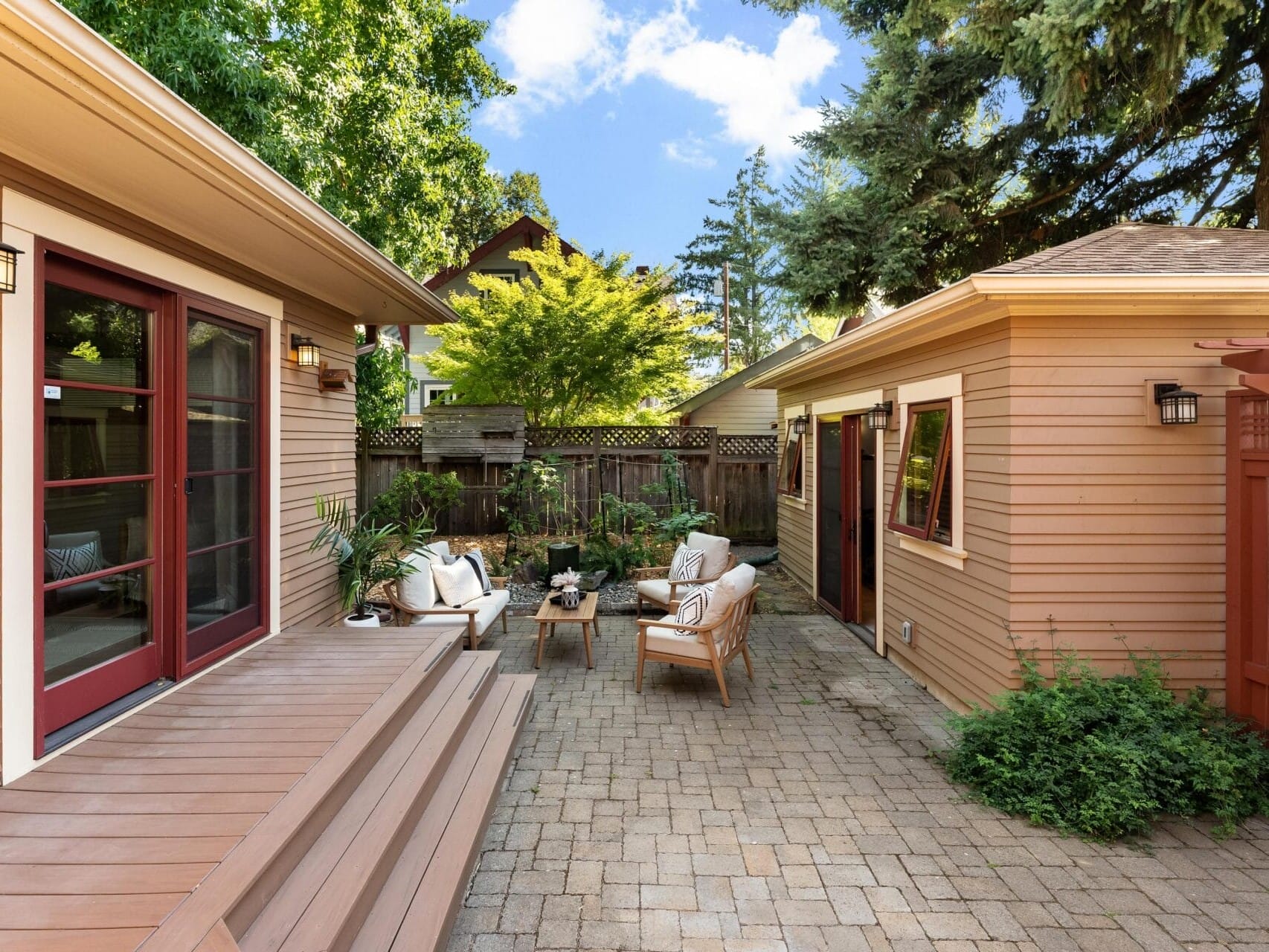 A cozy Portland, Oregon backyard patio features wooden furniture, including a sofa and armchairs arranged around a coffee table. The area is surrounded by a wooden fence and lush green trees, crafting a tranquil outdoor space under the city's iconic clear blue sky.