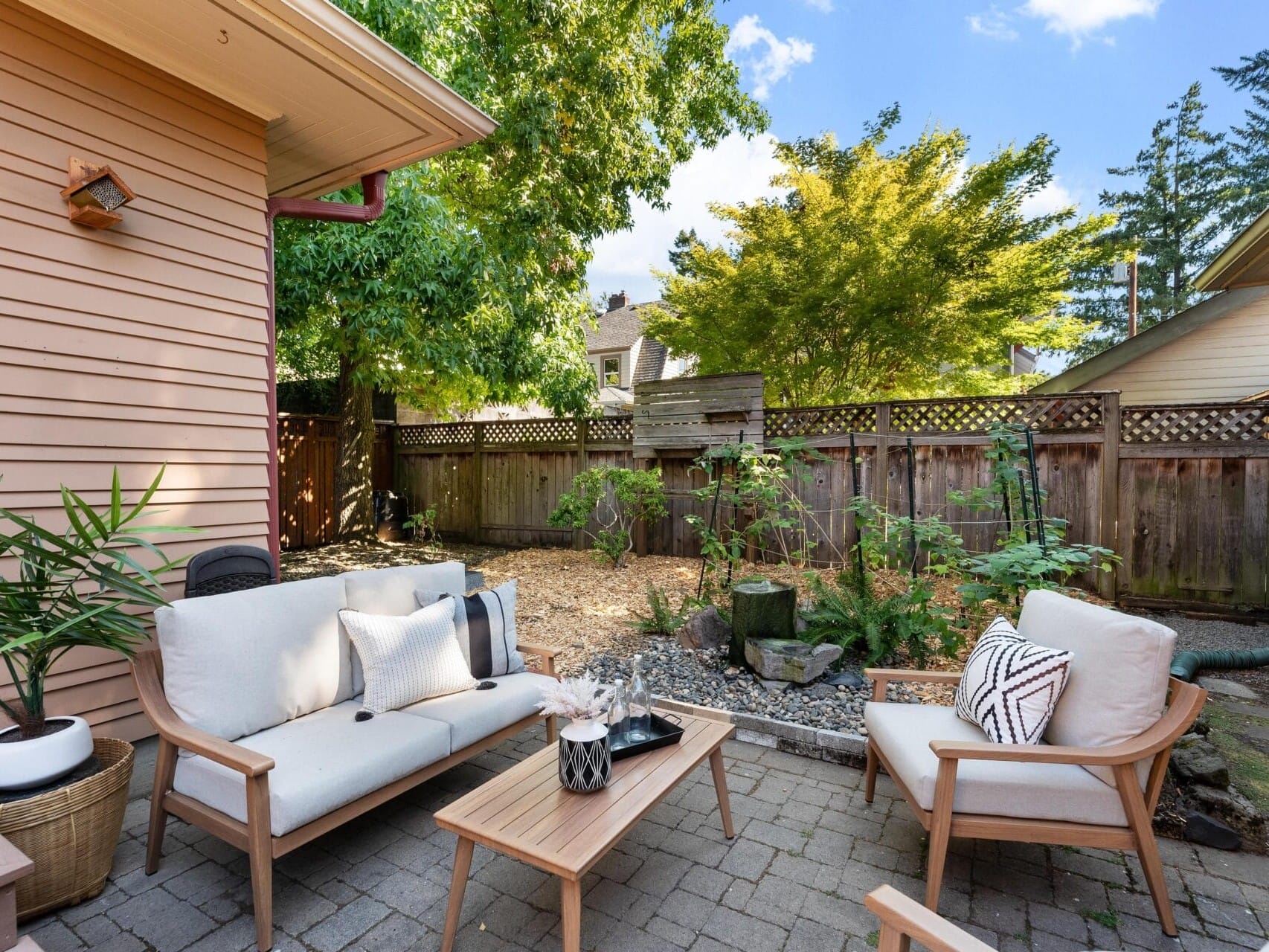 A cozy outdoor patio in Portland, Oregon features light-colored cushioned chairs and a wooden table on a brick floor, surrounded by greenery and a wooden fence. The area is shaded by trees, bathed in natural light, capturing the city's serene charm.