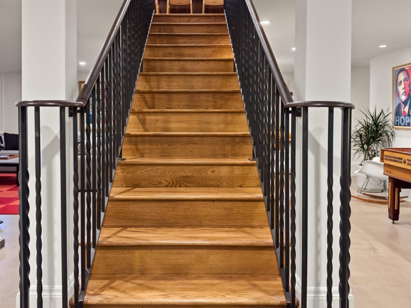 A wooden staircase with black metal railings leads to an upper floor in this modern Portland, Oregon, home. The room features white walls, a pool table, a plant, and a poster on the wall—an exquisite find in Portland real estate.