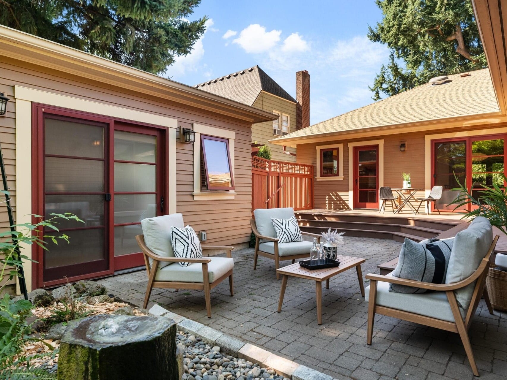 A sunlit patio in Portland, Oregon, features modern wooden chairs with white cushions around a small coffee table. Potted plants and decorative stones add charm, surrounded by beige buildings with red-trimmed windows and doors. A staircase leads to a raised deck.