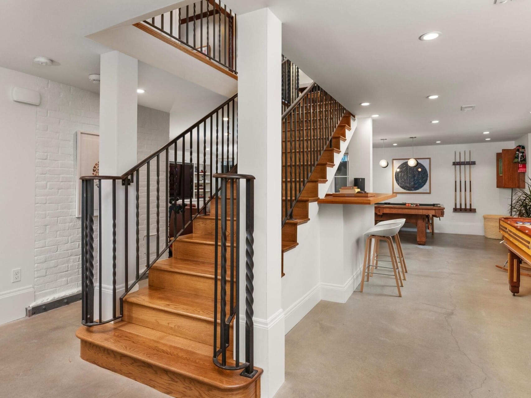 A modern basement in an upscale Portland Oregon real estate listing boasts wooden stairs, a pool table, and bar stools. With a polished concrete floor and artistic décor, this stylish space includes a stairway railing and a cozy seating area in the background, ideal for entertaining or relaxation.