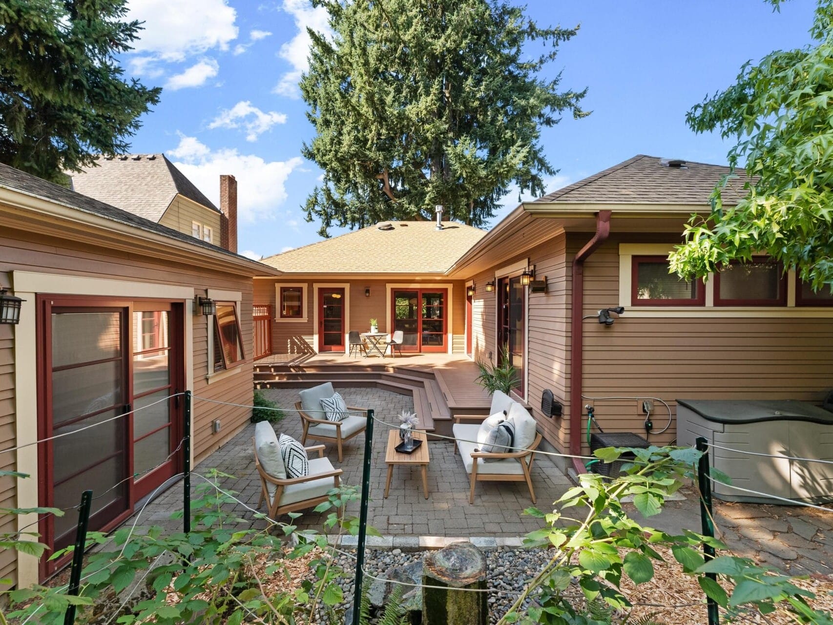 A cozy backyard patio in Portland, Oregon features wooden chairs and a table, surrounded by a wooden deck and two single-story buildings. Tall trees and a clear blue sky complete the serene setting.