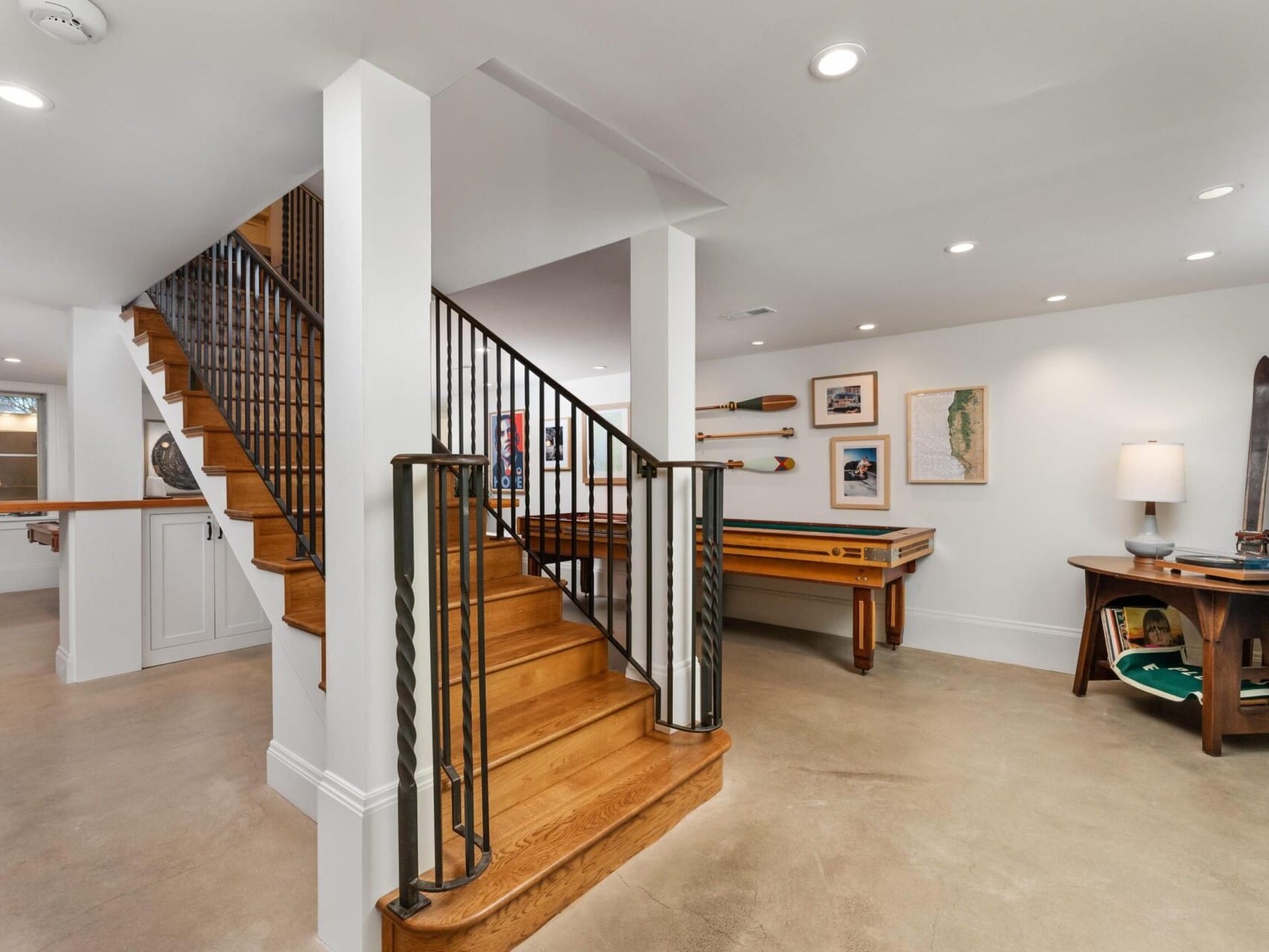 A spacious basement recreation room in Portland Oregon Real Estate features wooden stairs, a billiards table, and wall art with paddles and maps. The room boasts concrete flooring, a round wooden table with a lamp, and modern recessed lighting.