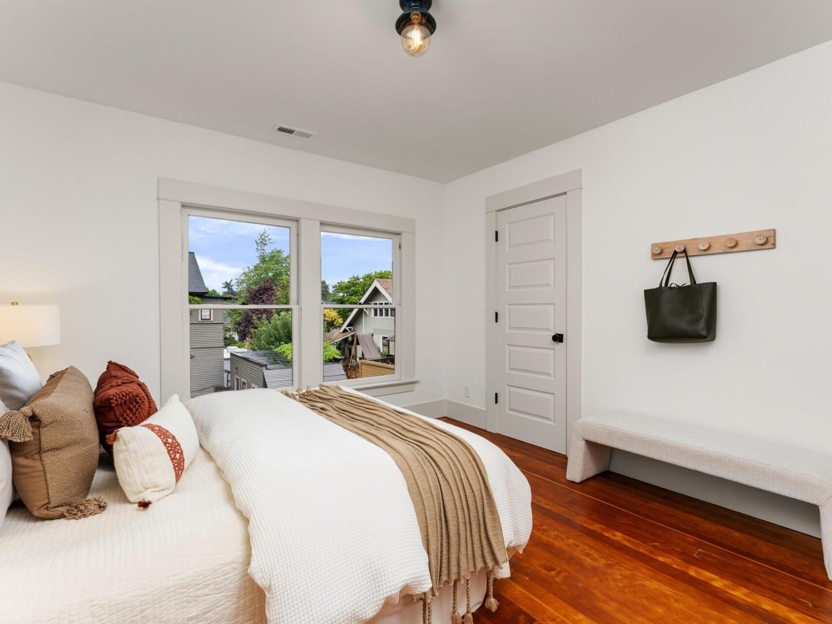 A cozy bedroom in Portland, Oregon features a large bed adorned with pillows and a beige throw. A window offers a view of neighboring houses. Beside the white door, a wall-mounted coat rack holds a bag, while a minimalist bench sits on the wooden floor.