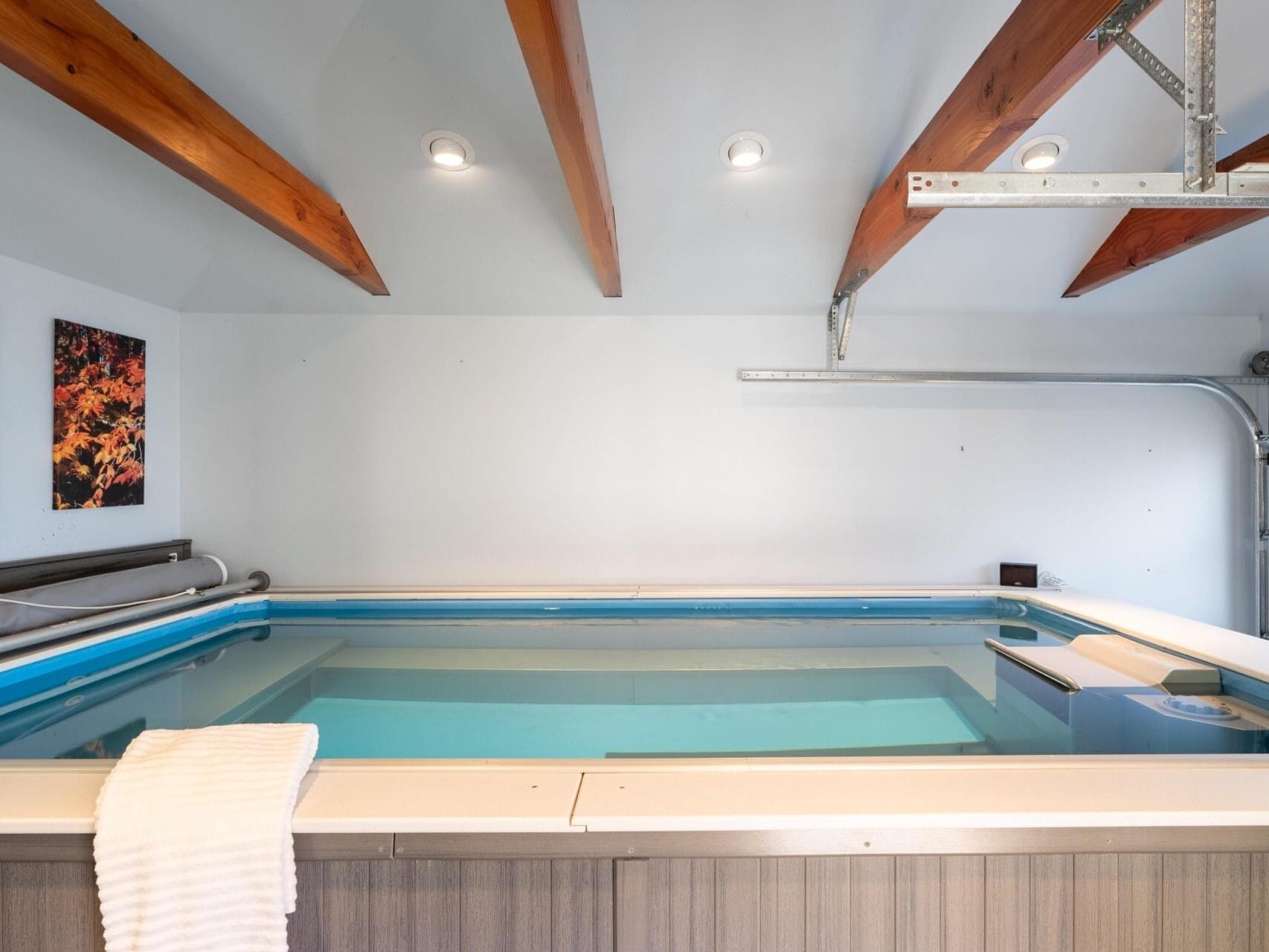 Indoor pool room in Portland, Oregon, featuring a rectangular pool with a towel draped over the edge. Wooden beams and recessed lights adorn the ceiling. Two abstract paintings are displayed on the wall, and a partially visible garage door is on the right.