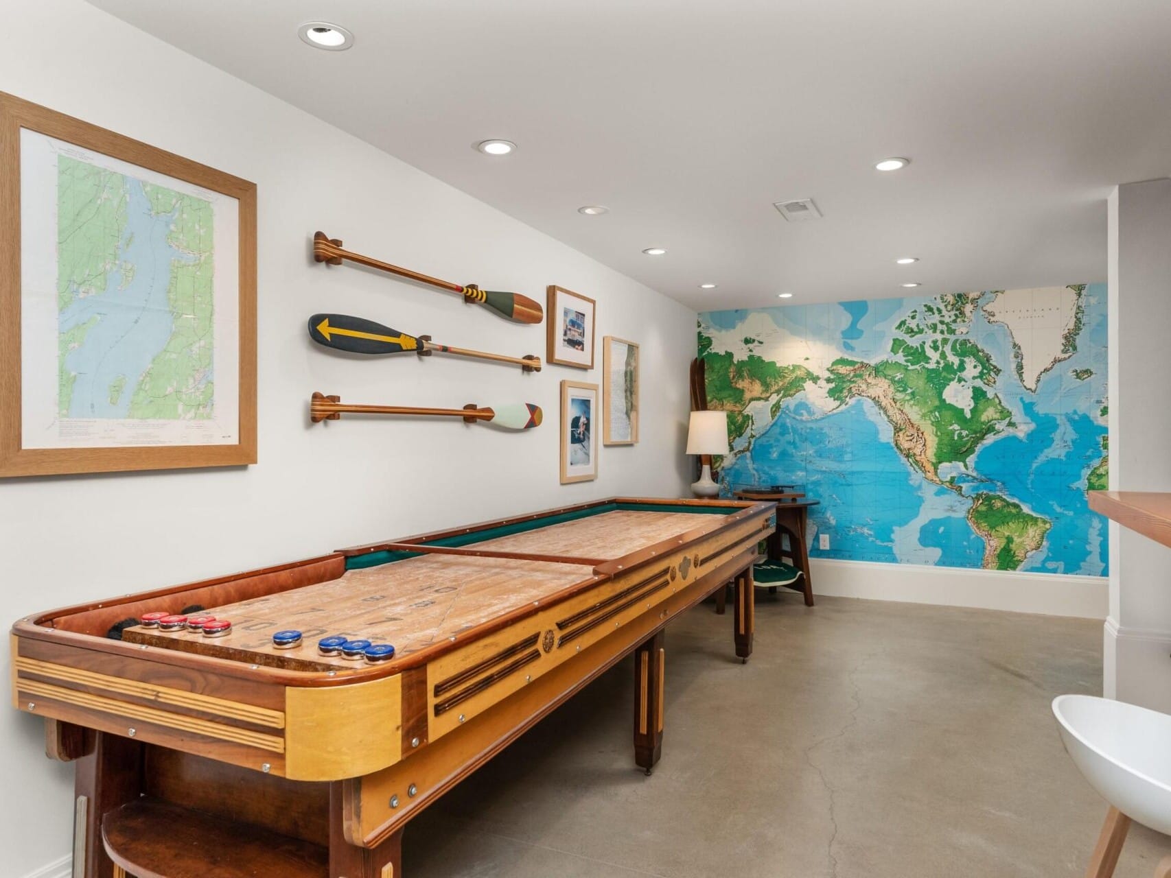 A room with a shuffleboard table, two framed maps, and a wall decorated with a large world map mural, exemplifying the style you'd expect from a top Portland realtor. Paddles and a lamp are visible in the corner. The floor is made of polished concrete.