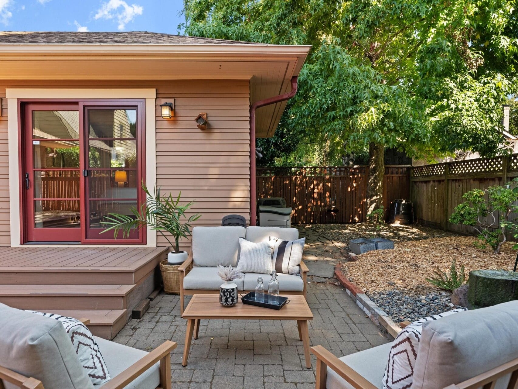 A cozy Portland, Oregon backyard patio with cushioned chairs and a wooden table set on a brick surface. The table holds a plant and glass pitcher. The beige house features red-framed sliding doors. Lush trees and a wooden fence complete the tranquil setting.