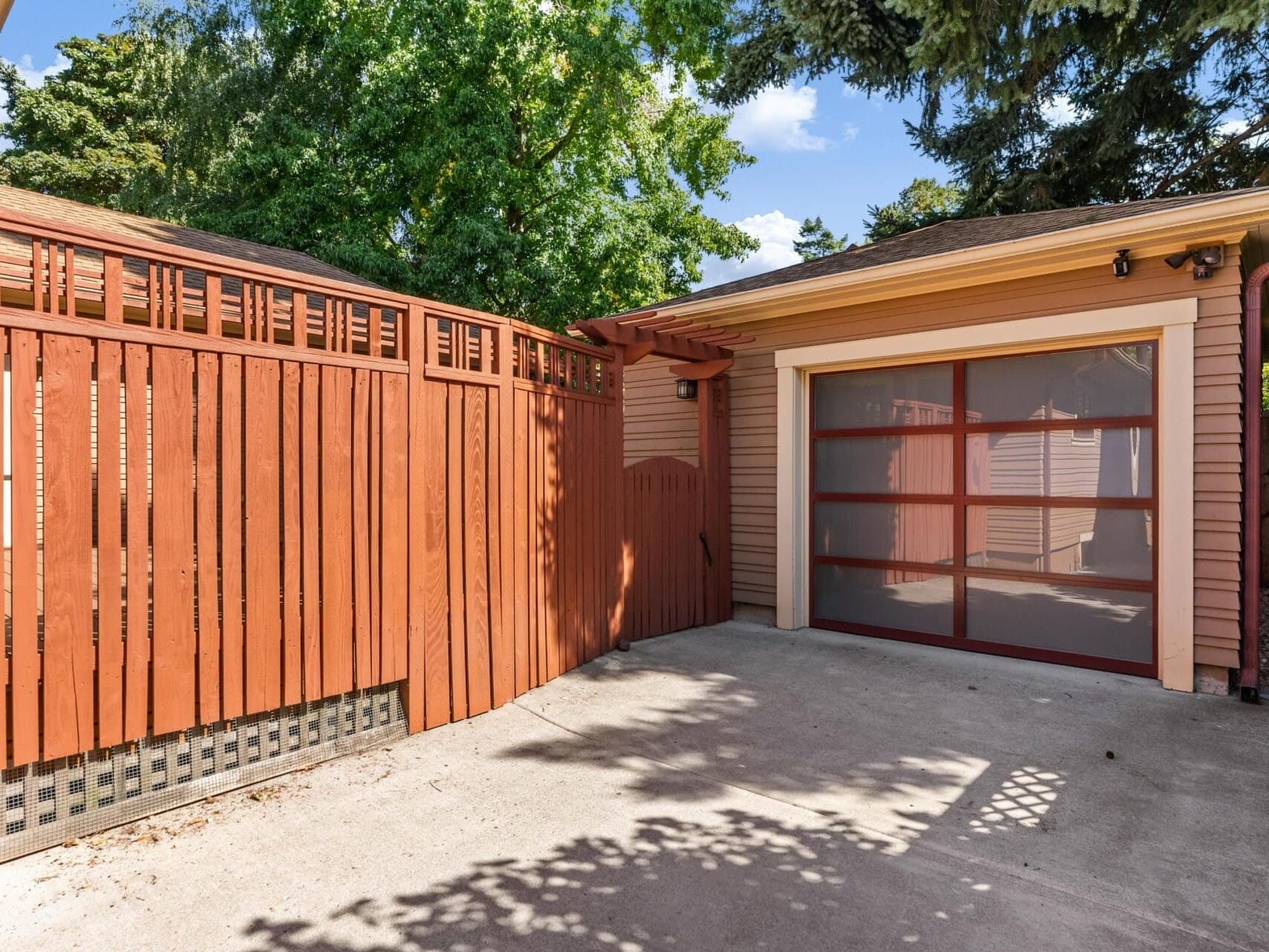 A modern garage in Portland, Oregon, boasts a wooden fence on the left and a driveway on the right beneath a clear blue sky. Surrounded by trees and sunlight, the garage door features glass panels.