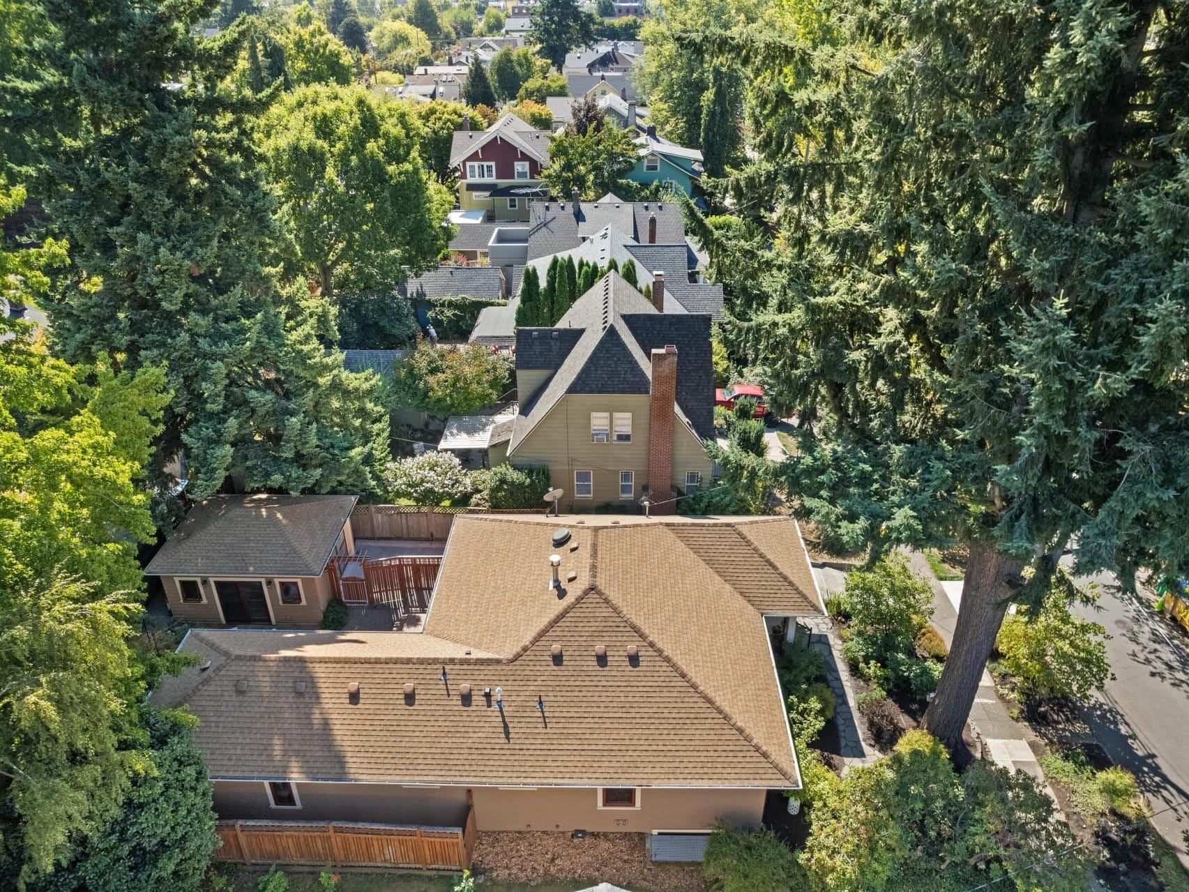 Aerial view of a residential neighborhood in Portland, Oregon, with houses surrounded by dense, tall trees. The brown-roofed house in the foreground is prominent, and other homes are visible in the background with a variety of roof styles and colors.