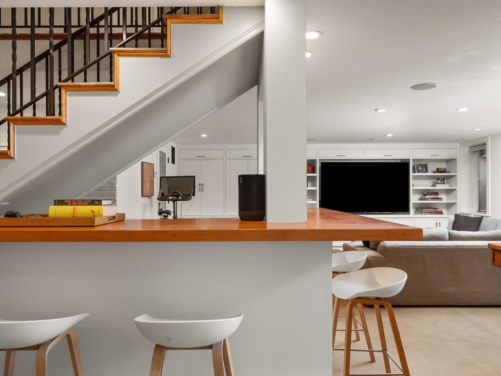 Modern living area featuring a wooden staircase, a bar with stools, and an open-plan lounge with a sectional sofa facing a TV. Books and decor items adorn the shelves, enhancing the cozy ambiance—a prime showcase of Portland Oregon real estate at its finest.