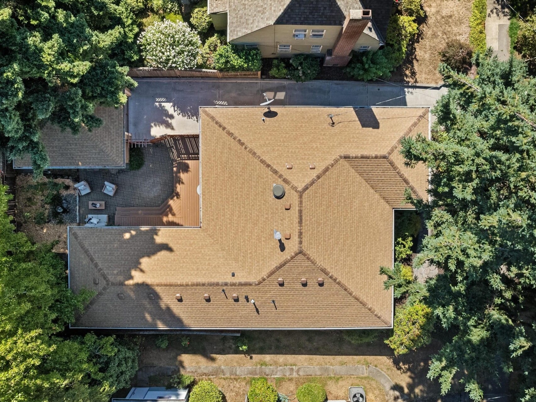 Aerial view of a beige, cross-shaped house with a tan roof surrounded by lush green trees. A fenced backyard with a patio area and outdoor furniture is visible. The house is adjacent to another building with a darker roof.