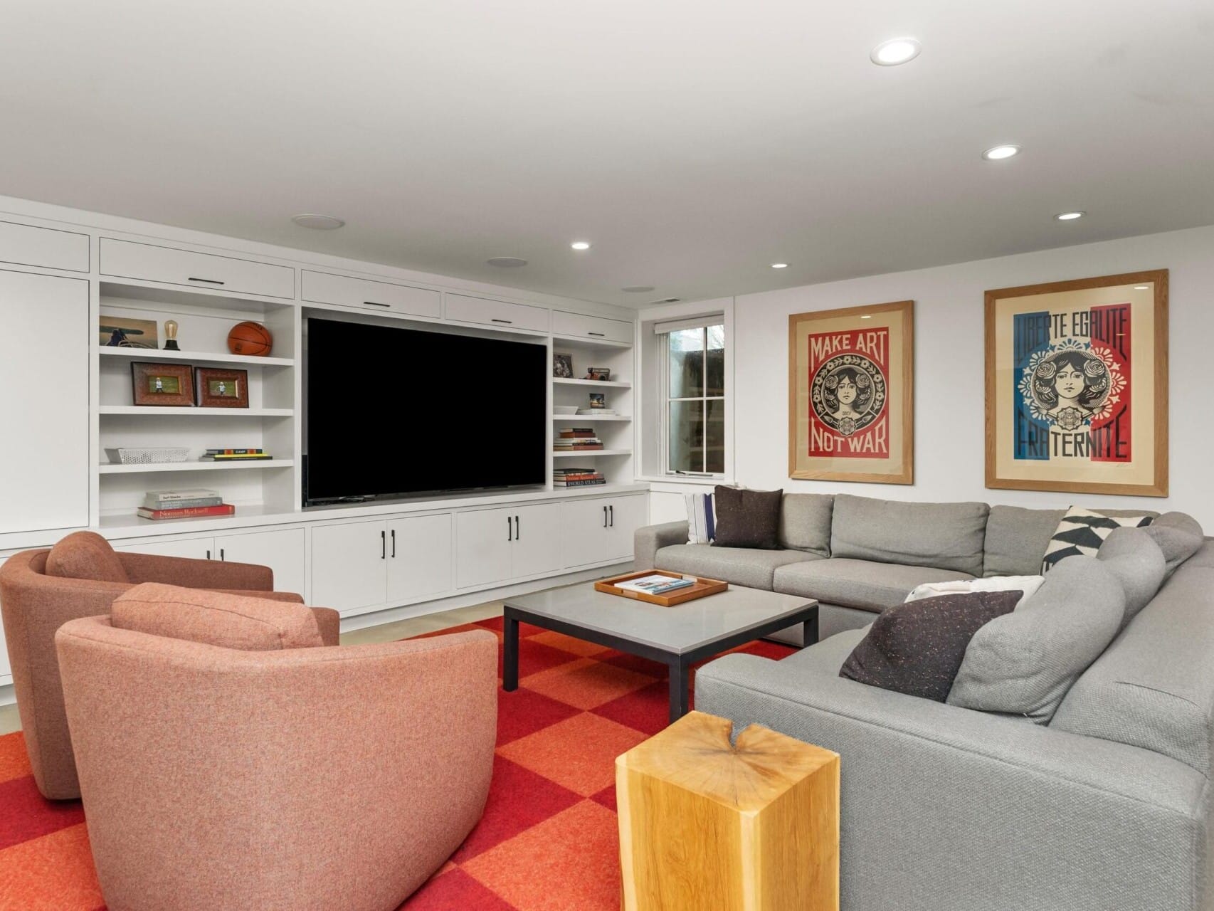 A modern living room in a Portland Oregon real estate gem features a large black screen on a built-in unit with white cabinets and shelves. There's a gray sectional sofa, two red chairs, and a black table on a red checkered rug. Two colorful framed artworks adorn the wall.