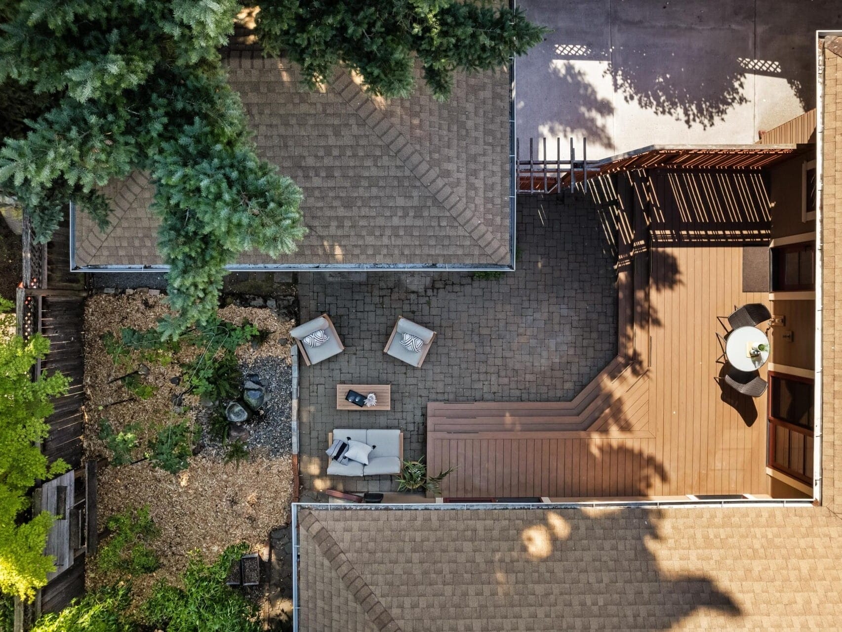 Aerial view of a wooden deck in Portland, Oregon, showcasing a person reclining on a sun lounger next to a coffee table and chairs. The deck is surrounded by buildings and trees, with a neatly arranged garden area on the left.