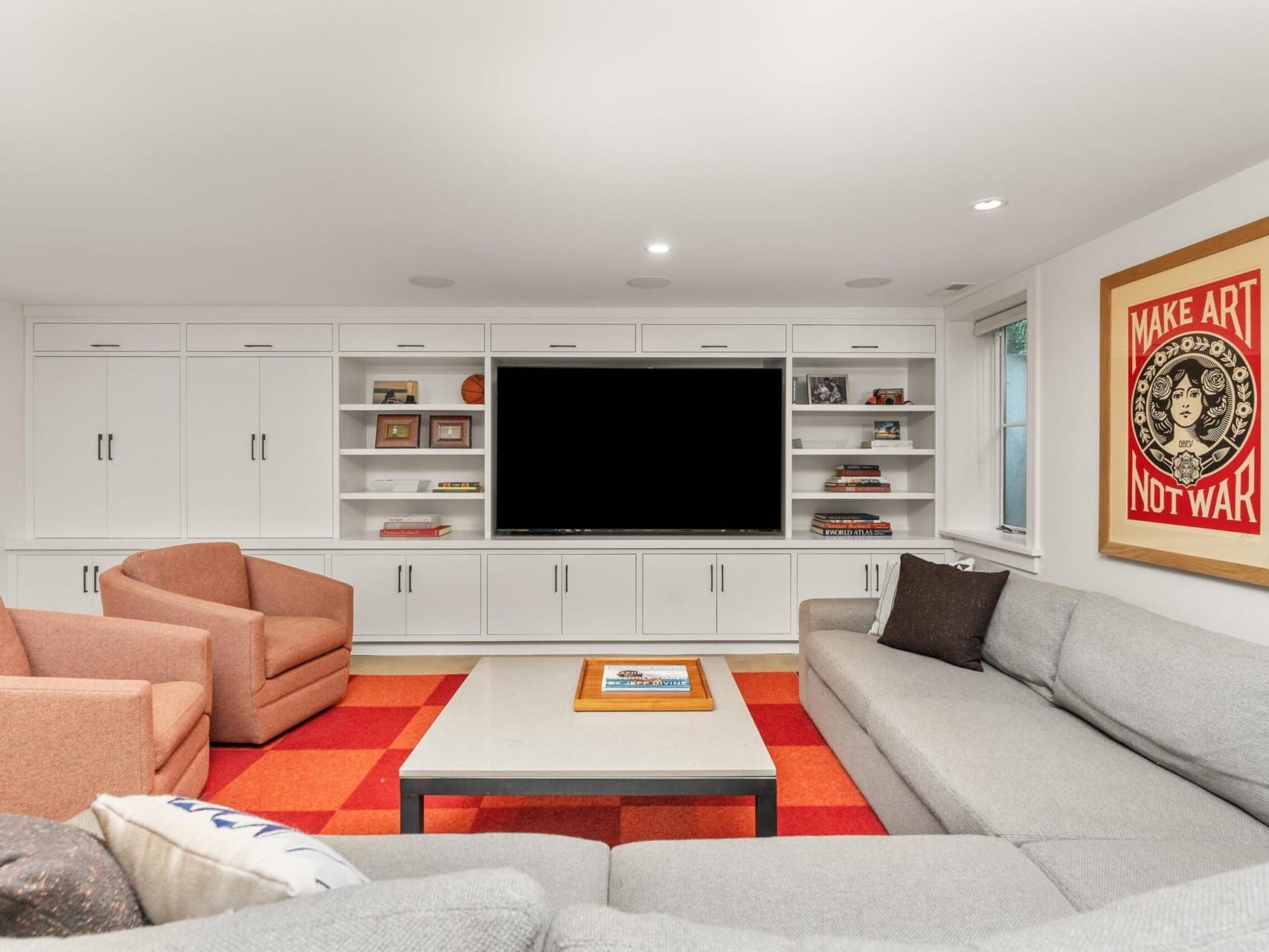 A modern living room in a Portland Oregon real estate gem features a large flat-screen TV surrounded by white cabinetry. Two brown armchairs and a gray sectional couch are arranged around a square coffee table on a red checkered rug. A poster on the wall reads, Make Art Not War.