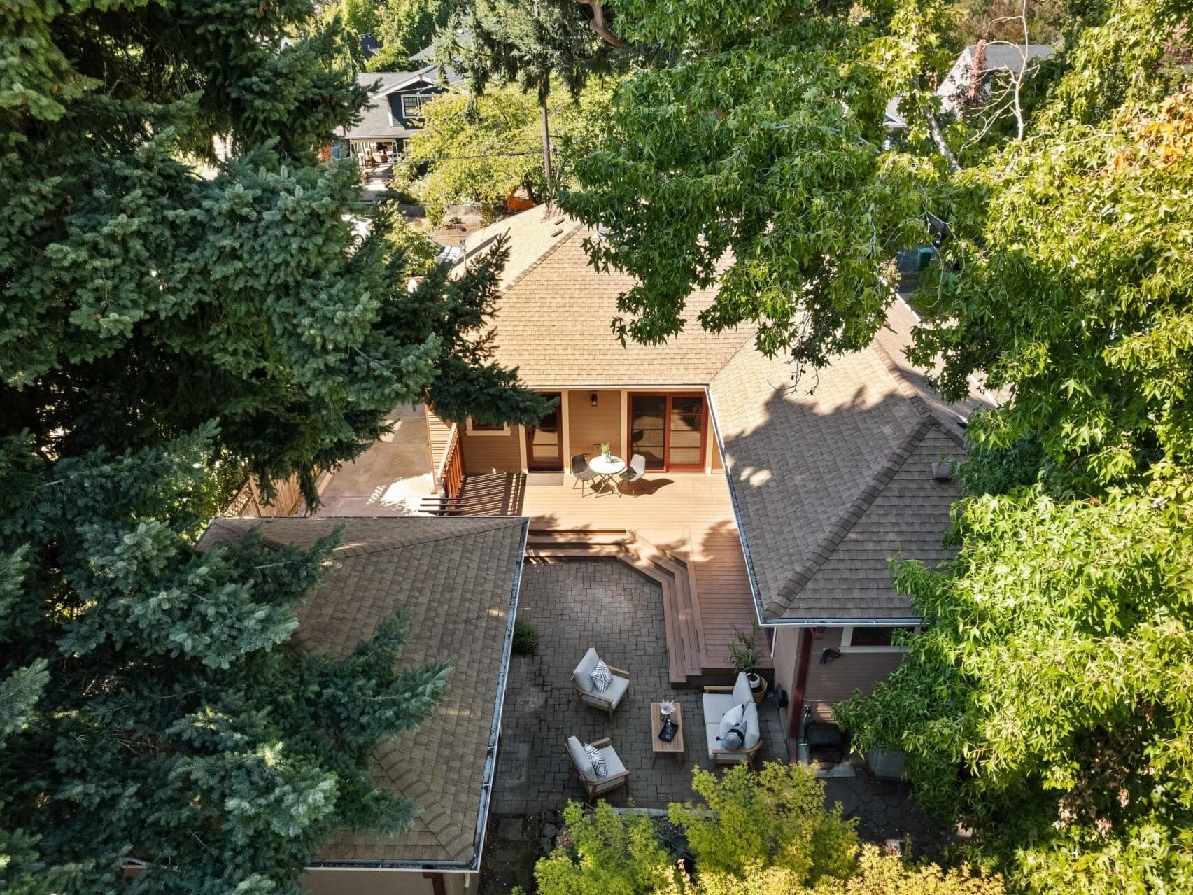 An aerial view captures a charming Portland, Oregon house nestled among lush green trees. It features a beige roof and a wooden deck adorned with outdoor furniture, including inviting chairs and a table.