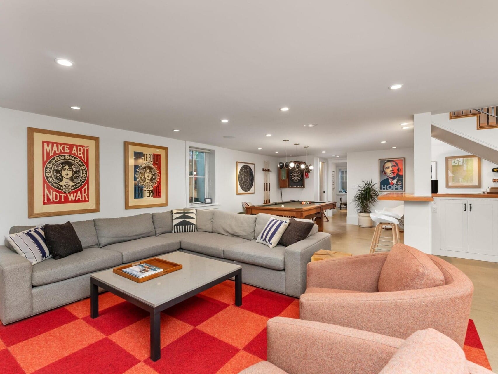 A modern living room featuring a large gray sectional sofa, two red armchairs, and a red checkered rug exudes Portland Oregon real estate charm. A pool table and framed artwork adorn the space, while a staircase with wooden steps elegantly completes this inviting ambiance.