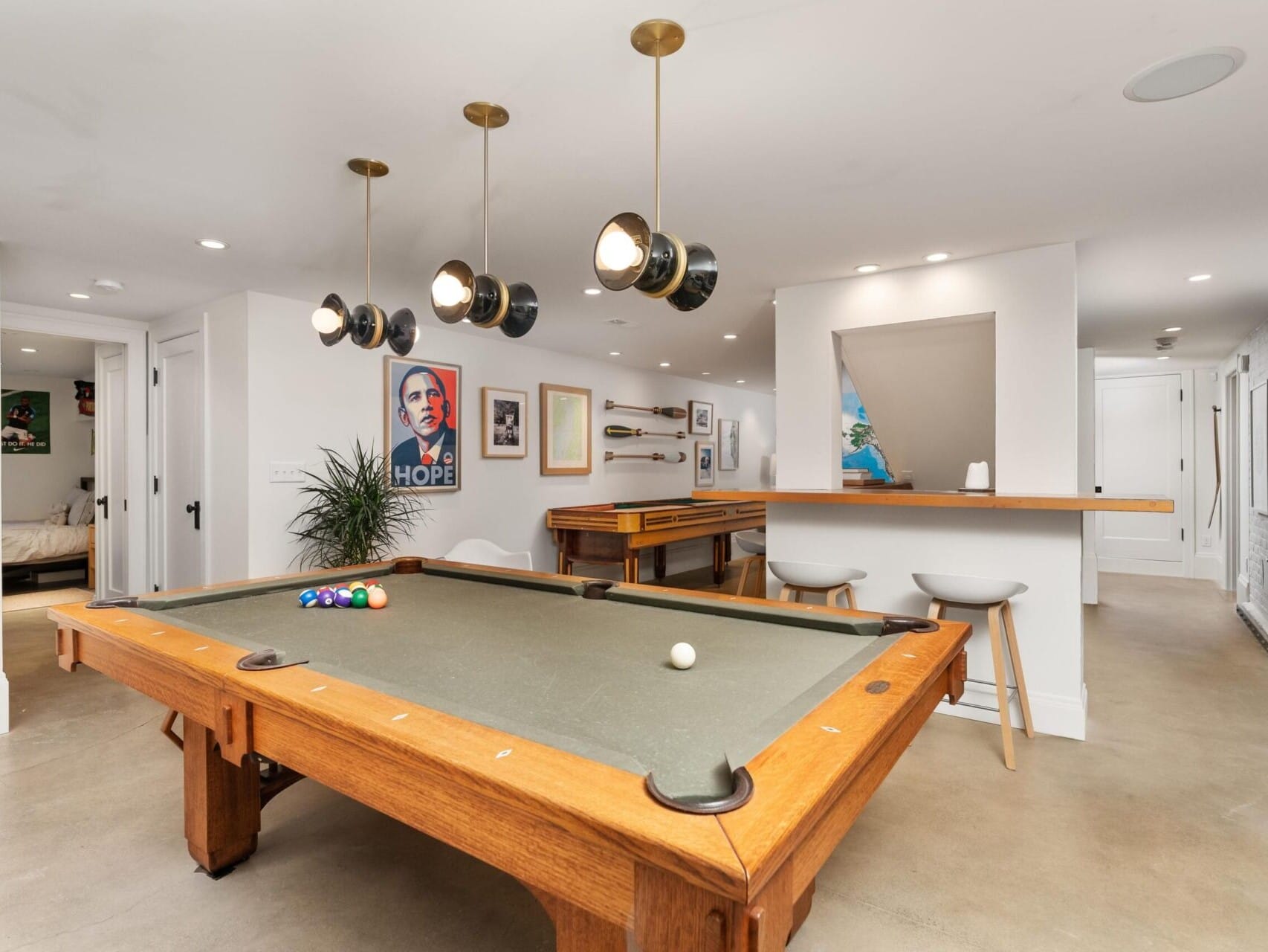 A recreation room showcases a pool table with a set of balls, stylish pendant lights, art and posters adorning the walls, and a chic bar area with stools. With a contemporary design and light-colored walls and flooring, it's an ideal highlight for any Portland Oregon real estate listing.