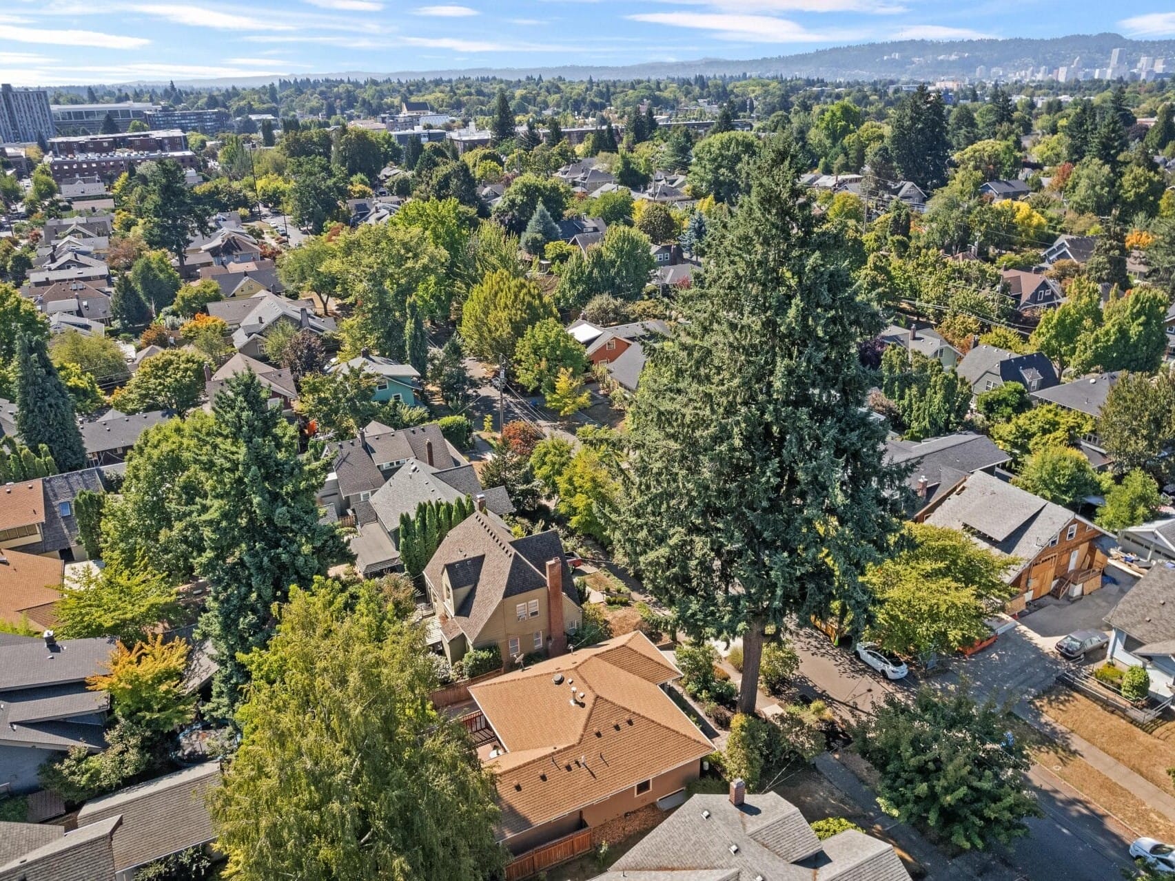 Aerial view of a lush suburban neighborhood in Portland, Oregon, filled with trees and houses. The homes are surrounded by greenery, showcasing a mix of residential architecture. The city skyline is visible in the distance under a clear blue sky.