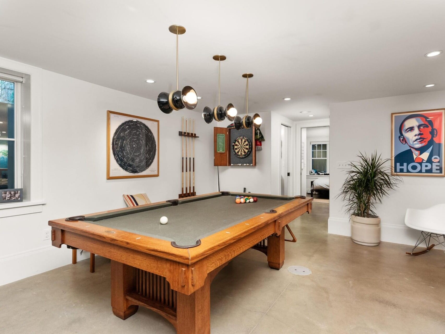 A modern game room, perfect for showcasing Portland Oregon Real Estate, features a pool table with billiard balls and cue stick, stylish wall art, and a dartboard. A plant graces the corner near a white rocking chair. Natural light filters through the window onto the polished concrete floor.