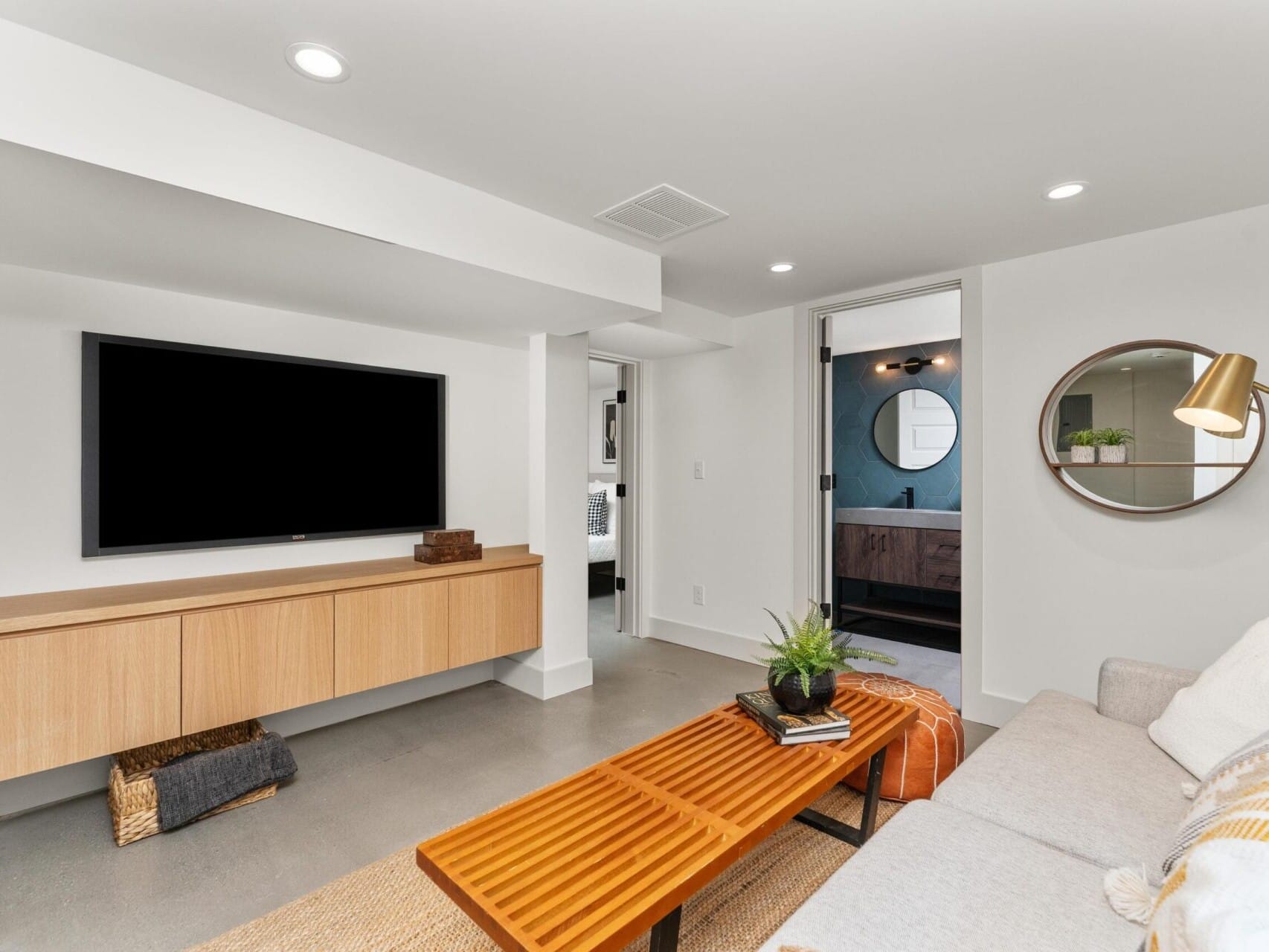 A modern living room in Portland, Oregon features a wall-mounted TV, wooden cabinet, and long coffee table. A beige sofa with cushions sits to the right. A doorway reveals a bathroom with a circular mirror. The space is well-lit with recessed lighting.