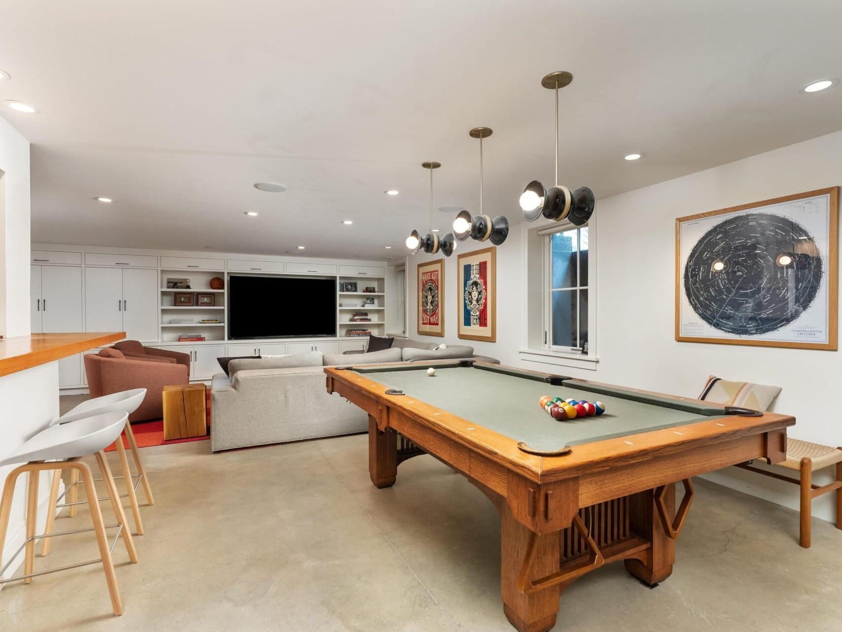 A modern recreation room designed for relaxation and entertainment, with a pool table in the foreground. The space, ideal for any Portland Oregon real estate gem, features a large TV, built-in shelves, cozy seating, and contemporary artwork. Neutral tones and stylish lighting create an inviting atmosphere.