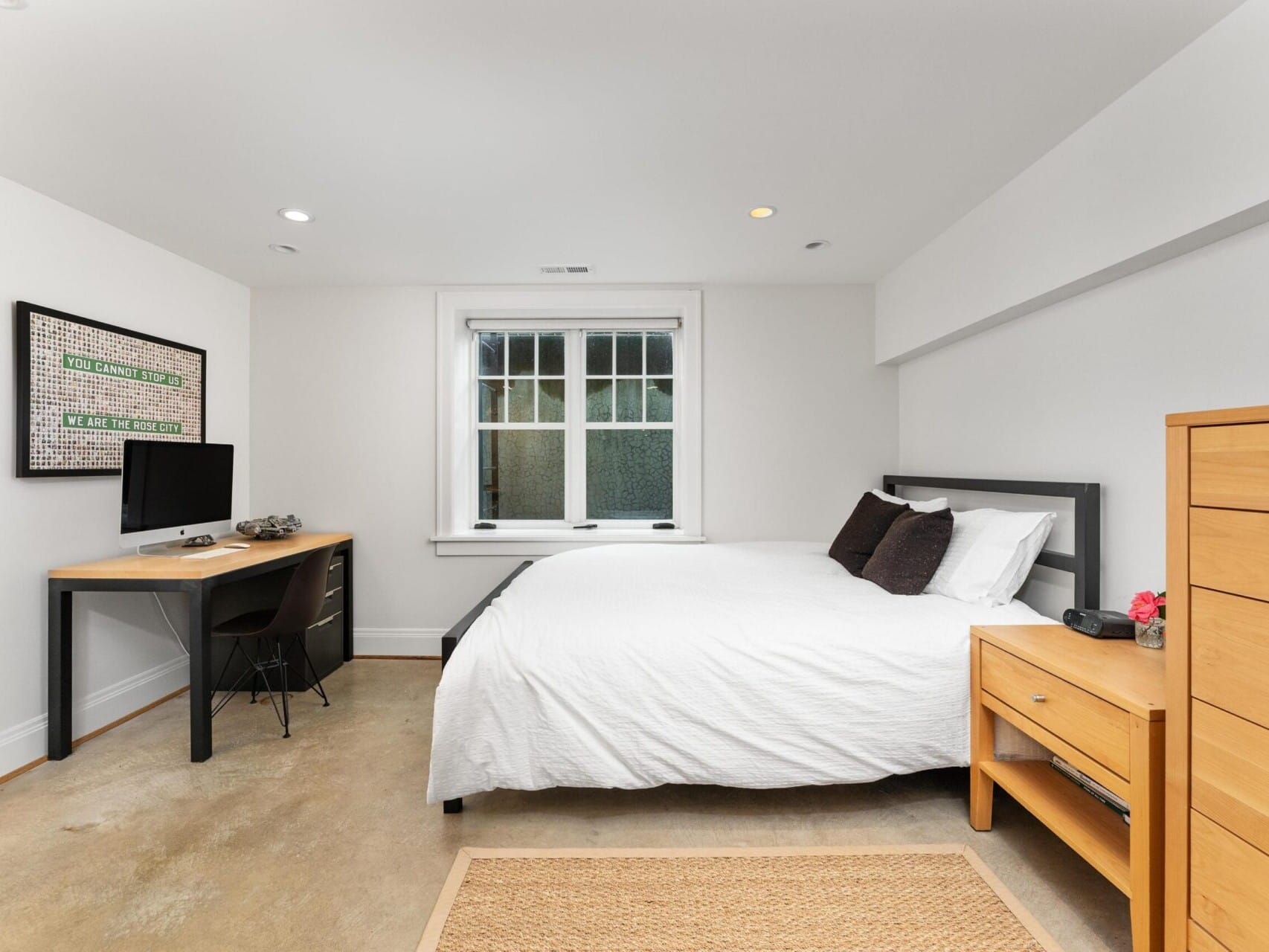 A neatly arranged bedroom in a Portland Oregon real estate listing features a bed with white linens and dark pillows. A wooden nightstand and dresser rest on the right, while a desk with a computer sits on the left. Light pours through a large window beneath a framed poster, creating an inviting space.