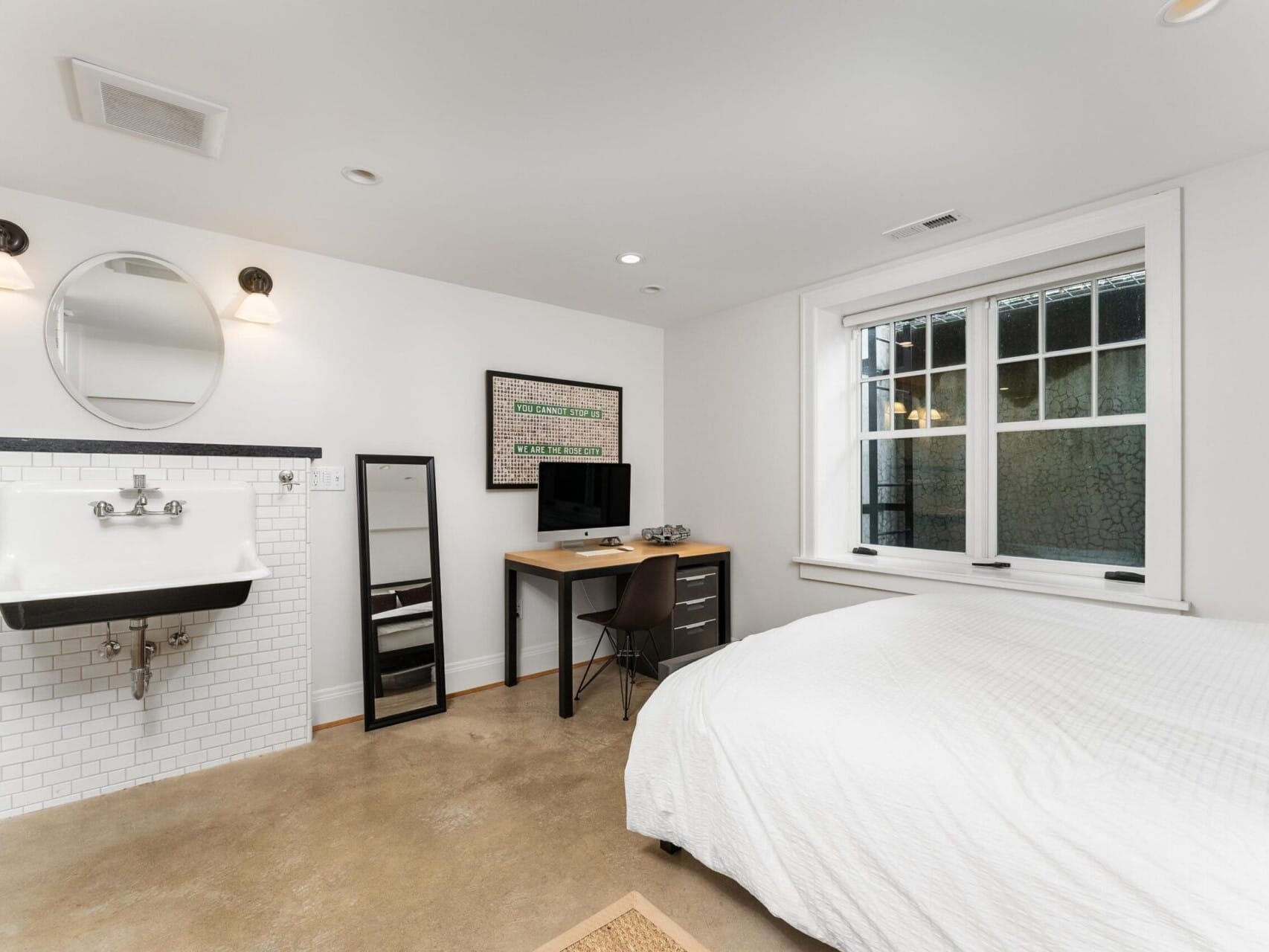 A minimalist bedroom in a chic Portland Oregon Real Estate listing features a bed, a wall-mounted sink with a round mirror, and a computer desk with chair. The room boasts white walls, expansive windows, and polished concrete floors, complete with trendy decor like the wall poster.