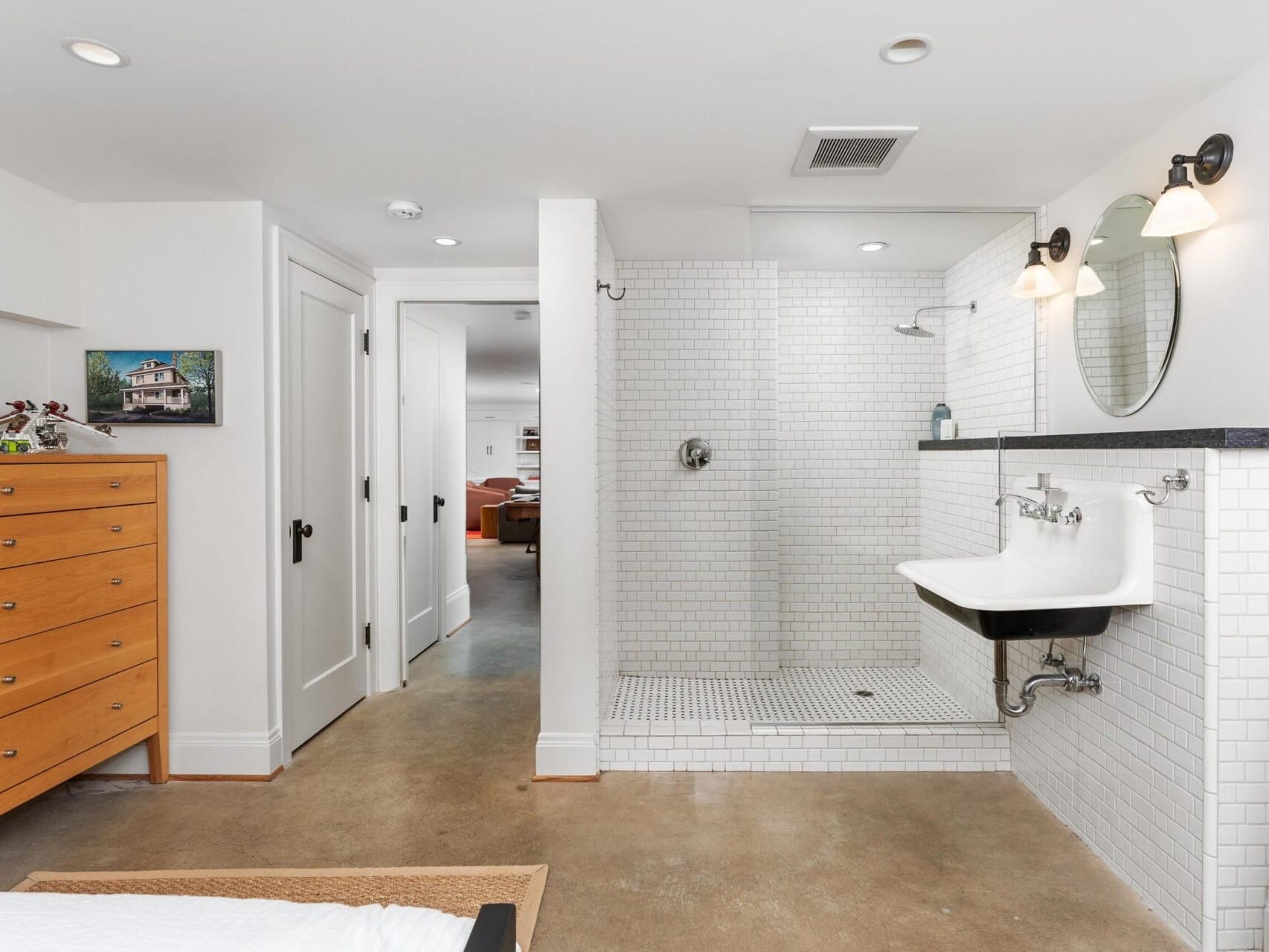 A modern bathroom in a Portland Oregon Real Estate listing showcases a large, open shower with white subway tiles, a wall-mounted sink, and dual sconces. A wooden dresser adds charm, while the polished concrete floor complements the view into another room through an open door.