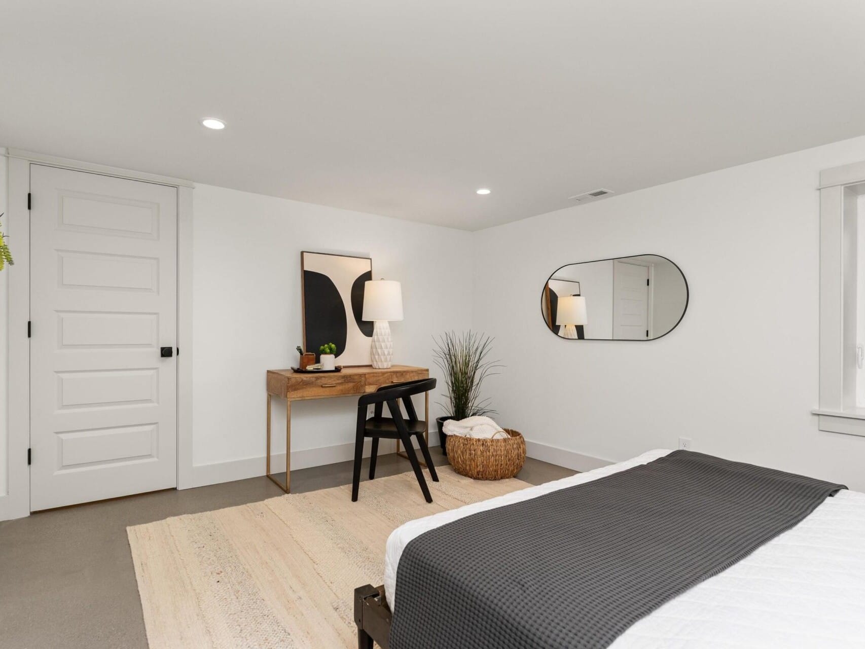 Minimalist bedroom in the heart of Portland, Oregon, featuring a bed with a gray blanket, a wooden desk with a chair, and a large oval mirror on the wall. Soft lighting from a lamp on the desk enhances the room's charm. A woven basket and plant adorn the corner against white walls and door.