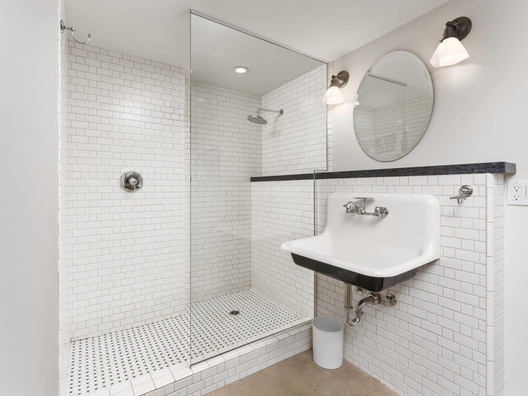 A minimalist bathroom, perfect for a Portland Oregon real estate listing, showcases white subway tile walls and a walk-in shower with glass enclosure. Black fixtures complement the white sink, topped by a round mirror with wall-mounted lights on each side. Ideal for any discerning Portland realtor client.