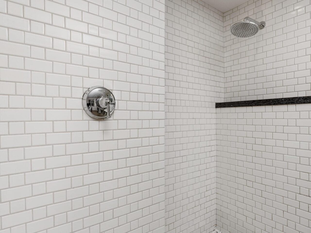 A modern shower with white subway tiles and a black accent strip, perfect for the discerning Portland Oregon real estate enthusiast. The chrome showerhead and control knob complement the basketweave tile floor, while a recessed ceiling light brightens this chic space.