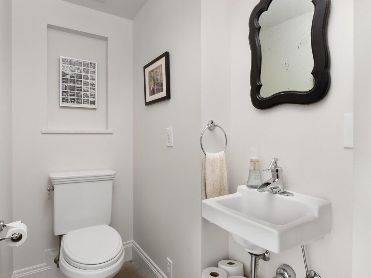 A small bathroom features a white toilet and sink, complemented by a mirror with a black ornate frame. A wall sconce light fixture above and a basket with toilet paper below add charm. With a small framed picture on the wall, this space showcases Portland Oregon real estate elegance.